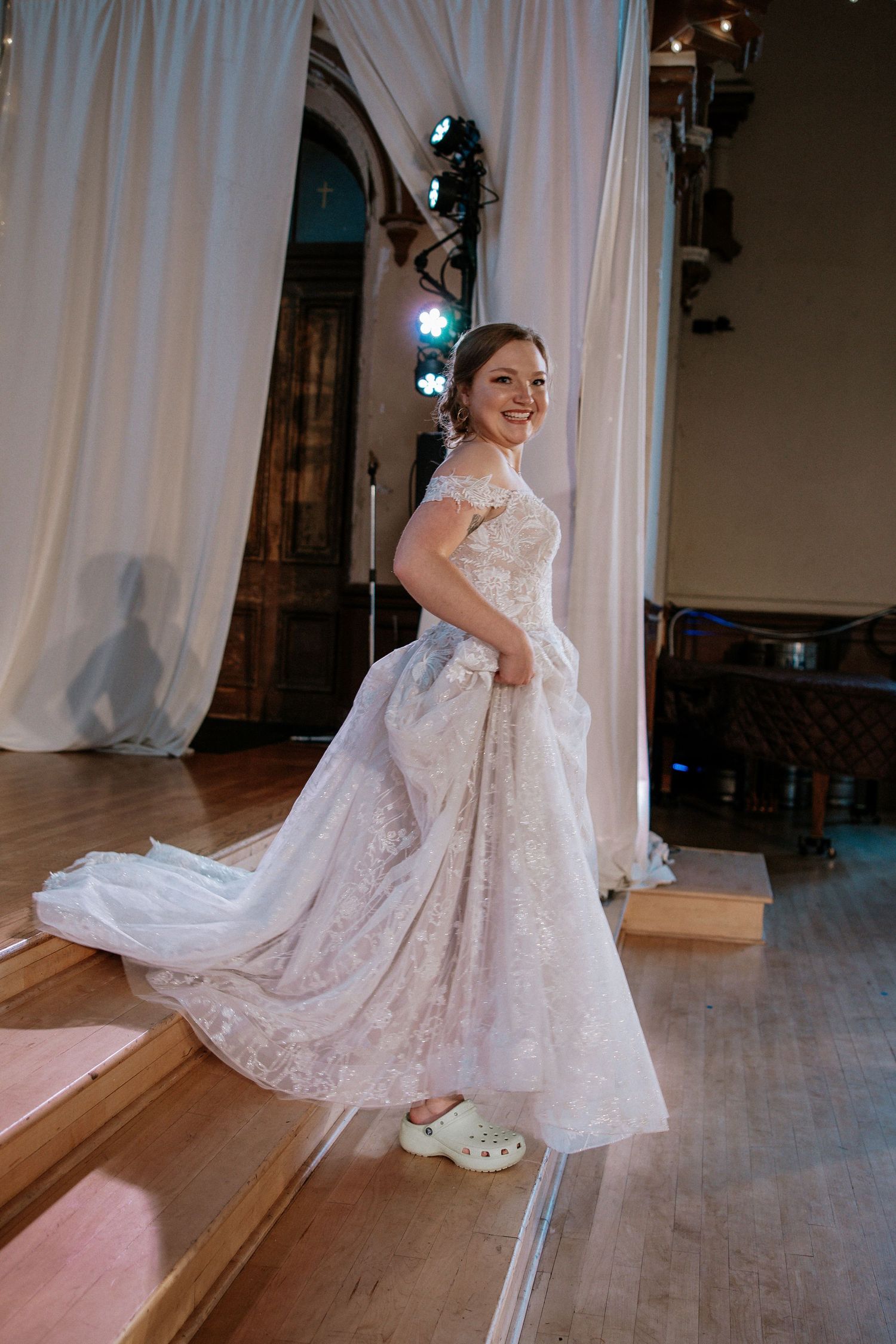 Smiling bride twirls in a stunning lace wedding gown wearing white Crocs on a wooden dance floor at Urban Artifact in Cincinnati, Ohio
