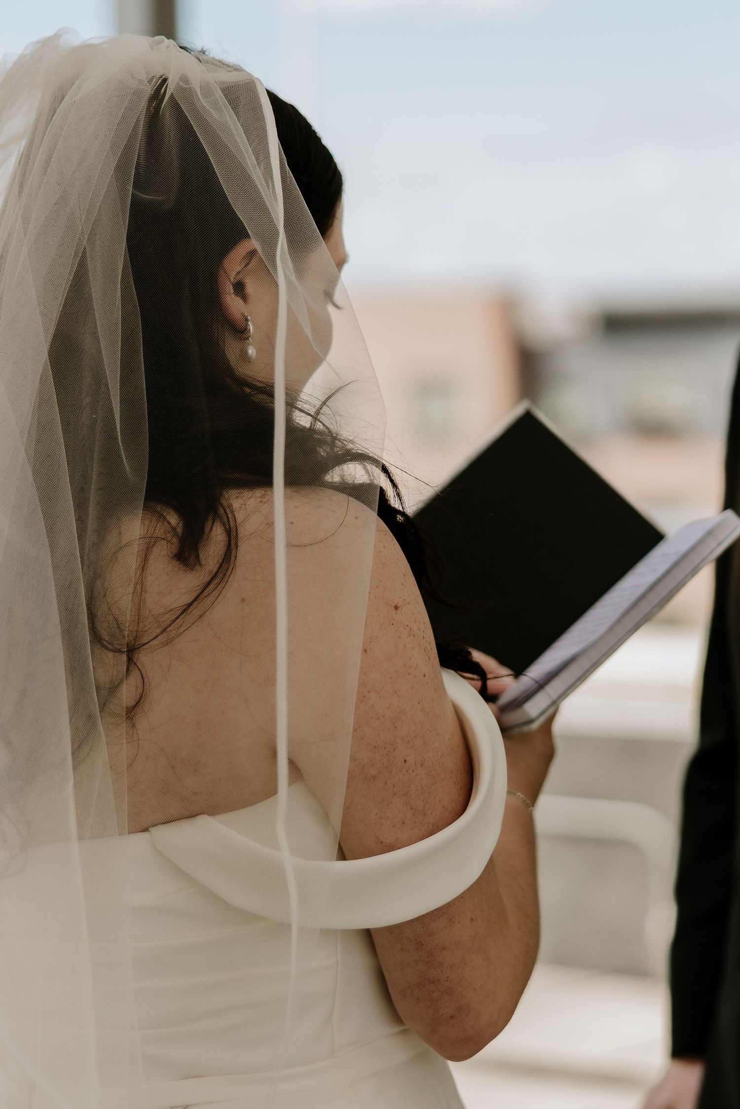 Bride in white off-shoulder gown and veil reads from a book during outdoor rooftop wedding ceremony.