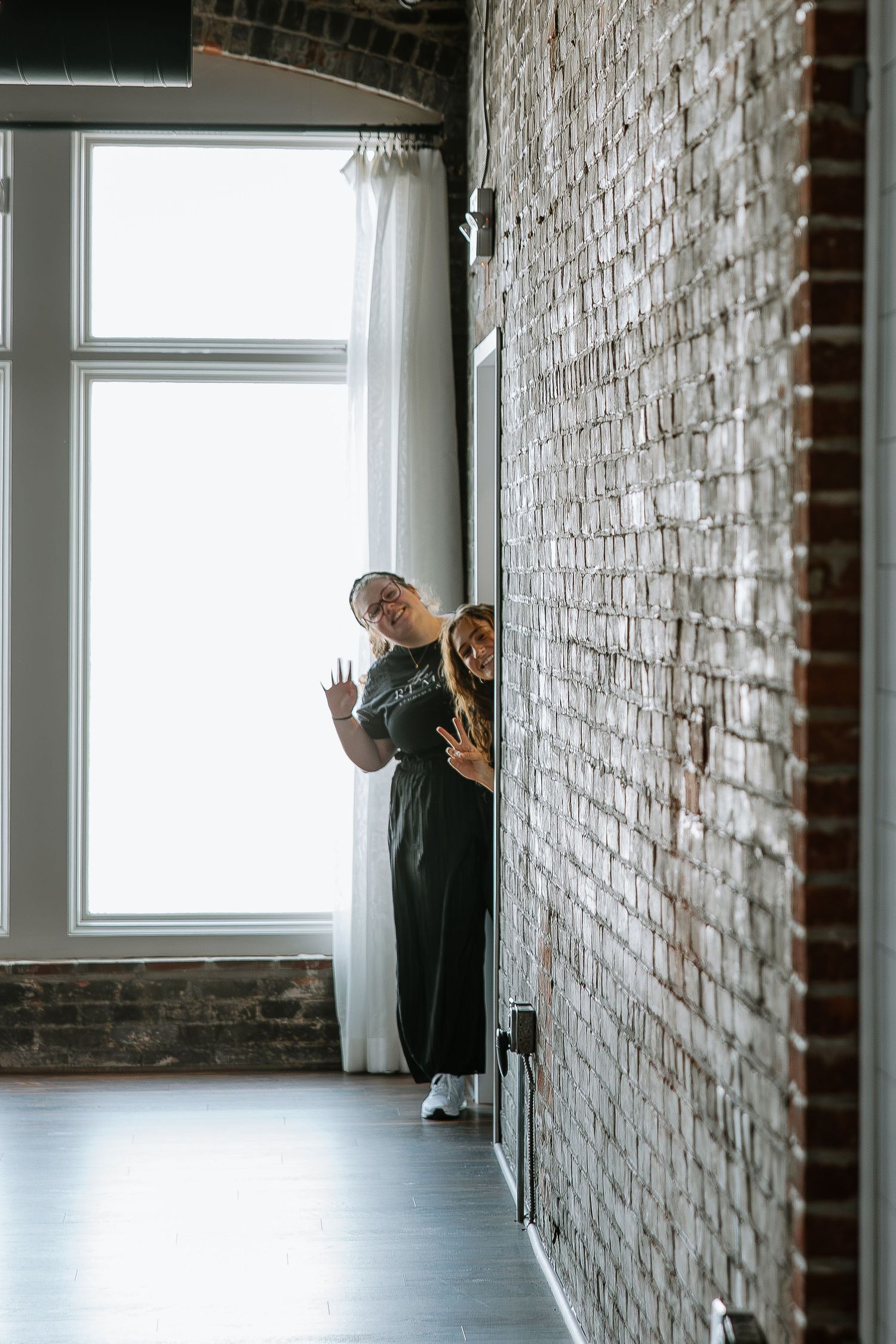 Two women peeking around a brick wall near a bright window at a wedding venue.