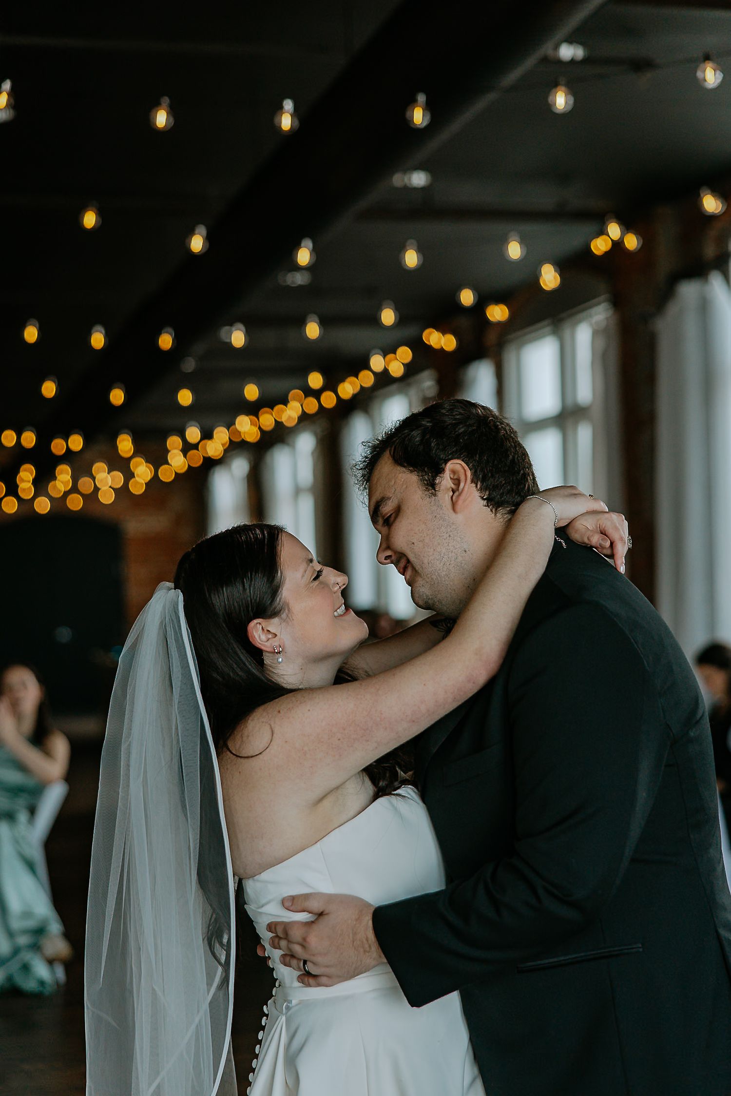 Bride and groom embrace tenderly during first dance under glowing string lights at wedding reception.