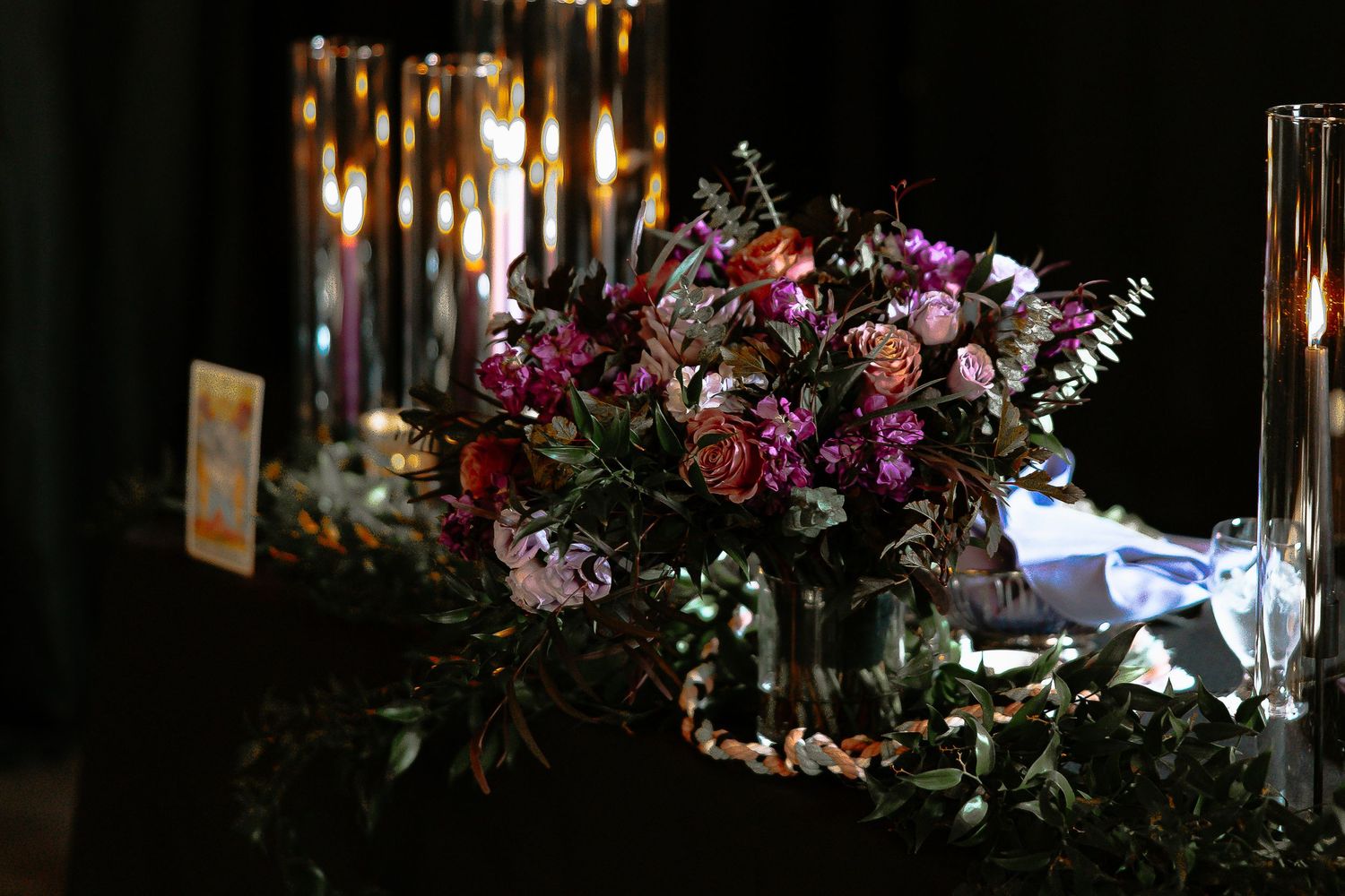 Lush dark floral centerpiece with purple and pink blooms surrounded by candles at a wedding reception.
