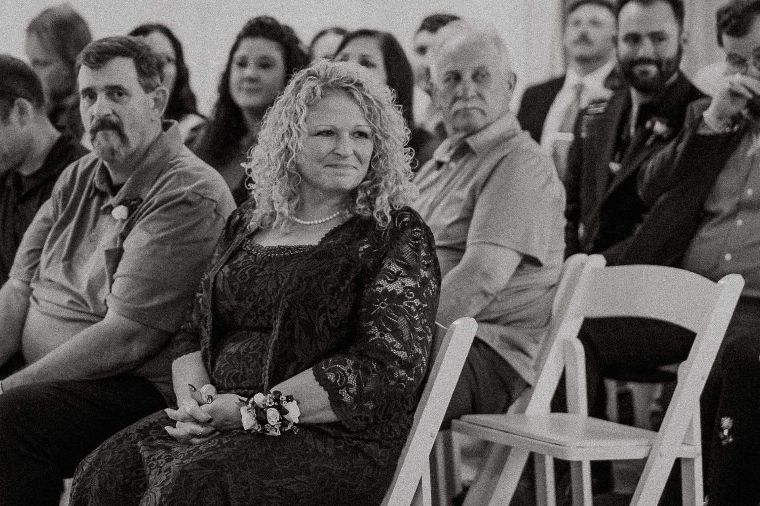 Black and white photo of curly-haired woman in lace dress seated at wedding ceremony.