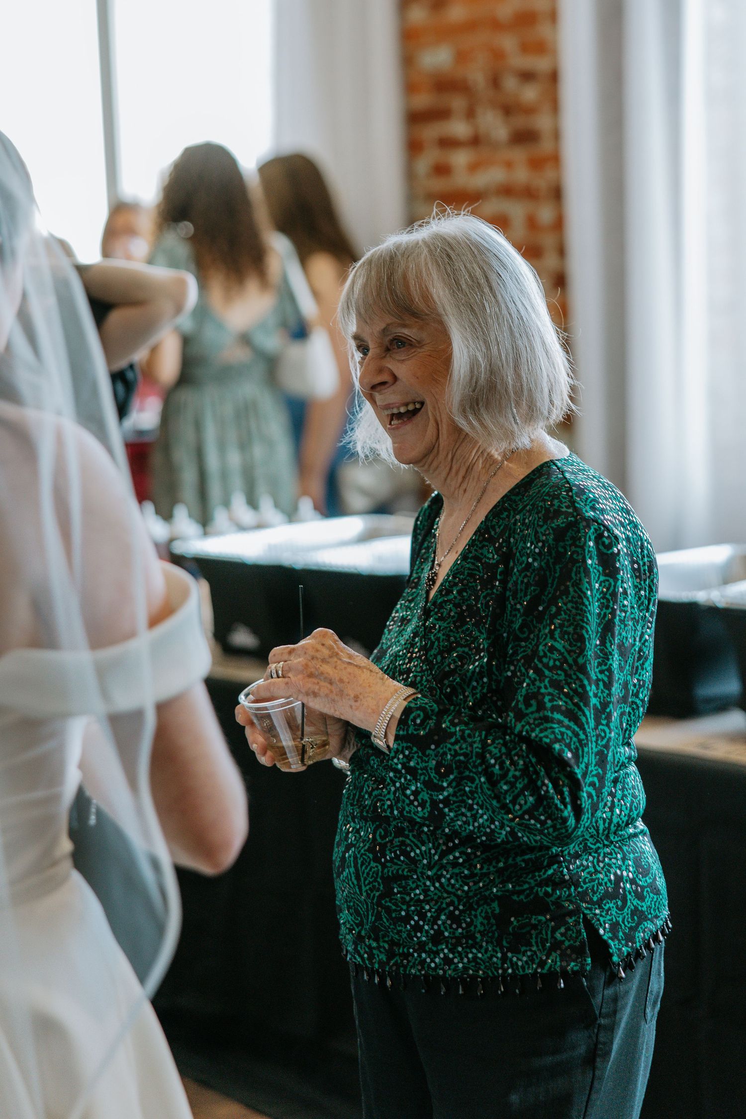 Elderly woman in green sequin top smiling and chatting with bride at wedding reception.