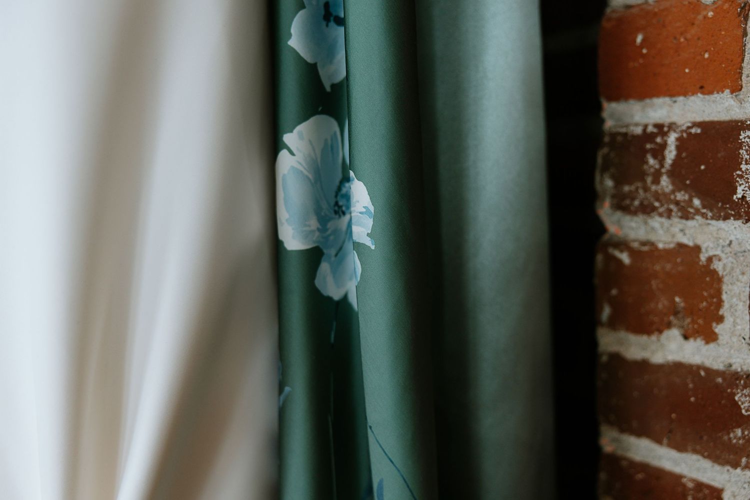 Green bridesmaid dress with delicate blue floral embroidery hanging beside white wedding gown near brick wall.