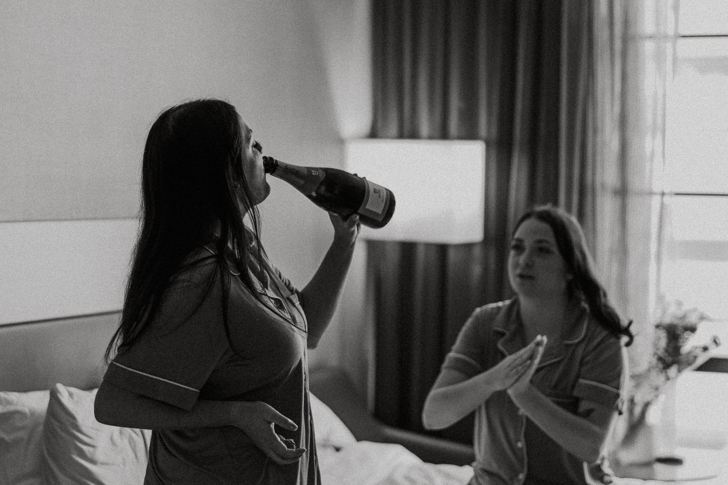 Black and white photo of bridesmaid drinking champagne from bottle in hotel room before wedding.