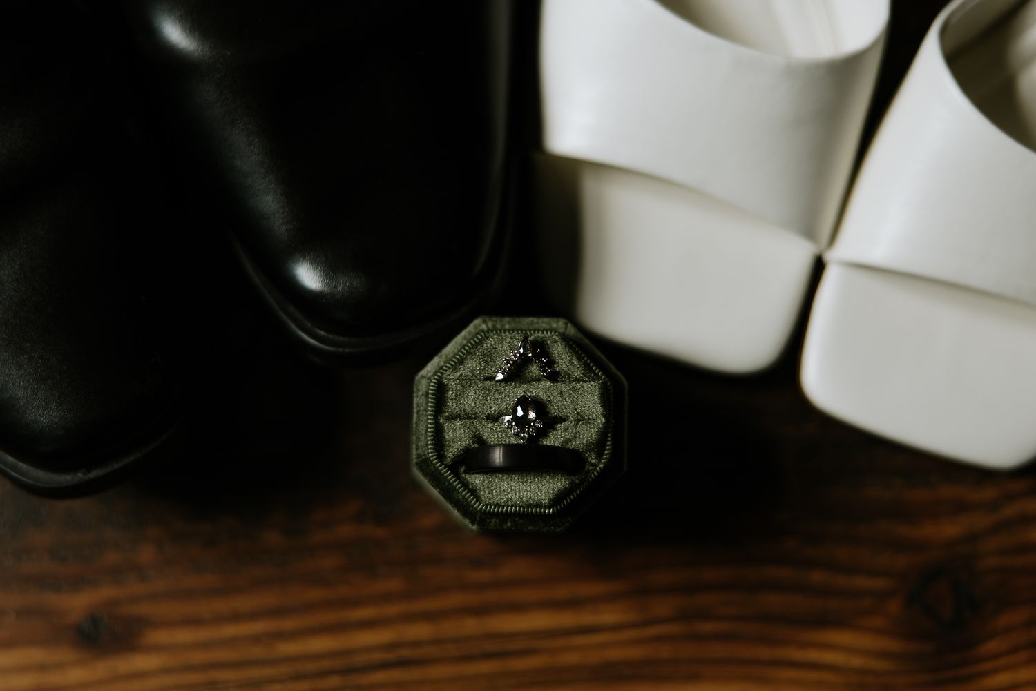 Wedding rings displayed in an ornate dark octagonal box between black dress shoes and white heels.