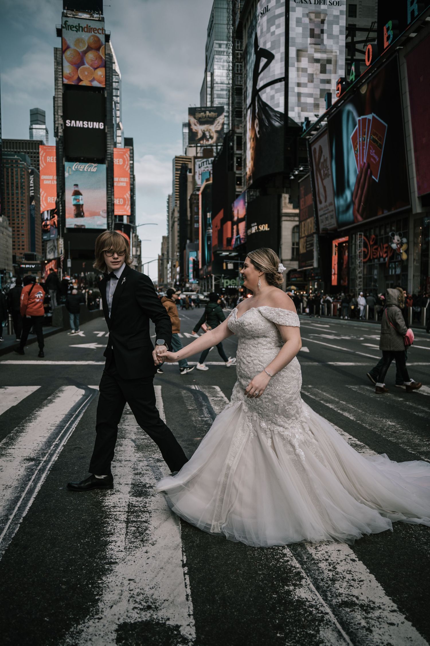 Bride in lace mermaid gown and groom in tuxedo walking hand in hand through Times Square NYC.