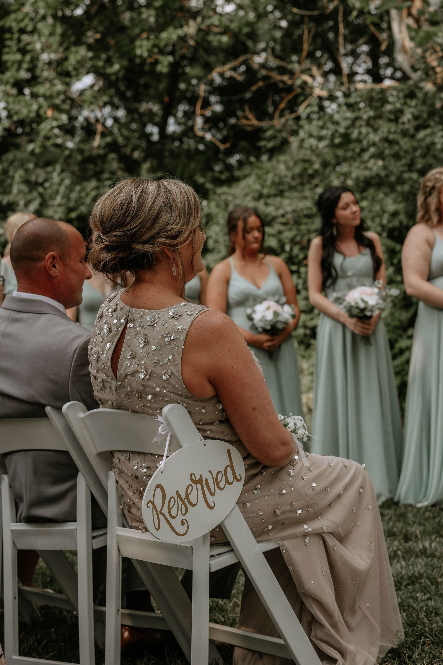 Woman in beaded gown sits in reserved chair at outdoor wedding ceremony with bridesmaids in sage green dresses.