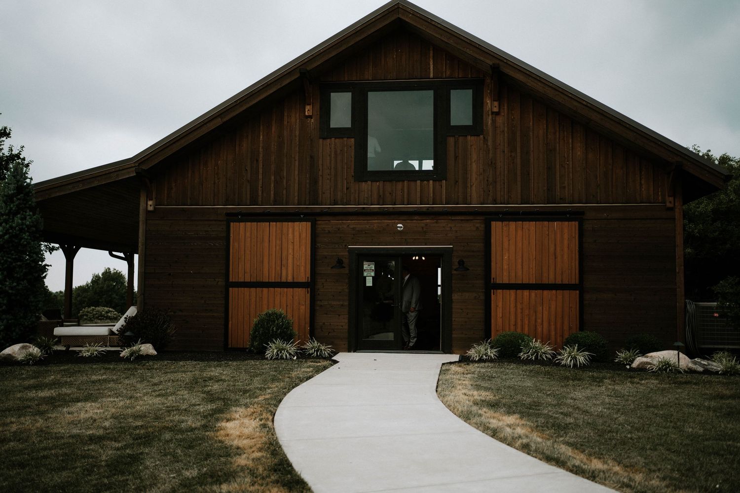 A rustic dark wood barn wedding venue with sliding doors and a concrete pathway leading to the entrance.