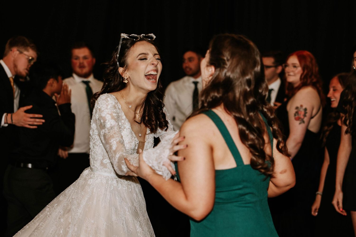 Bride in white lace gown laughing joyfully with bridesmaid in green dress during wedding reception dance.