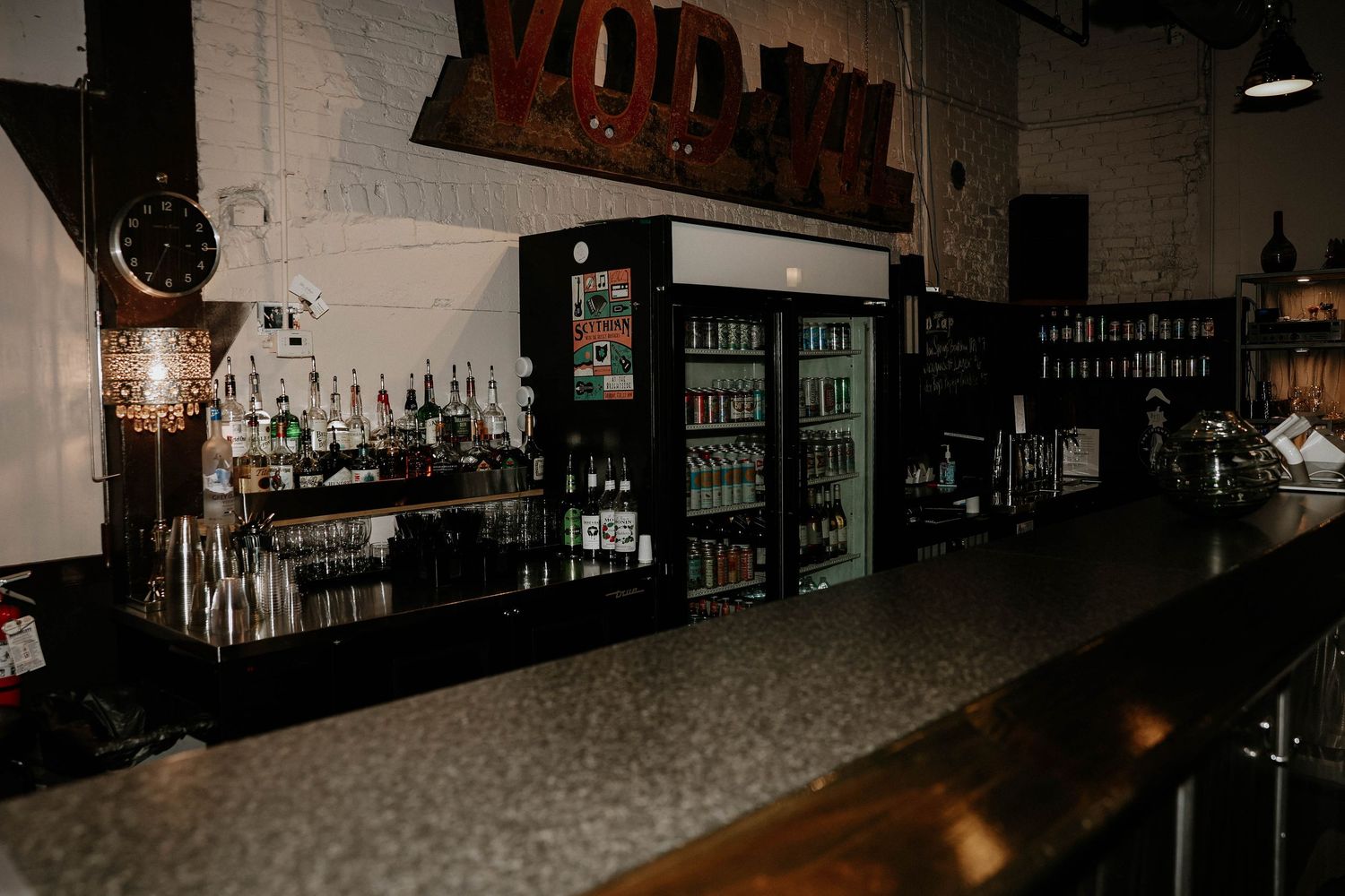 Dimly lit bar interior with a long counter, refrigerated drink displays, and shelves of liquor bottles against a brick wall.