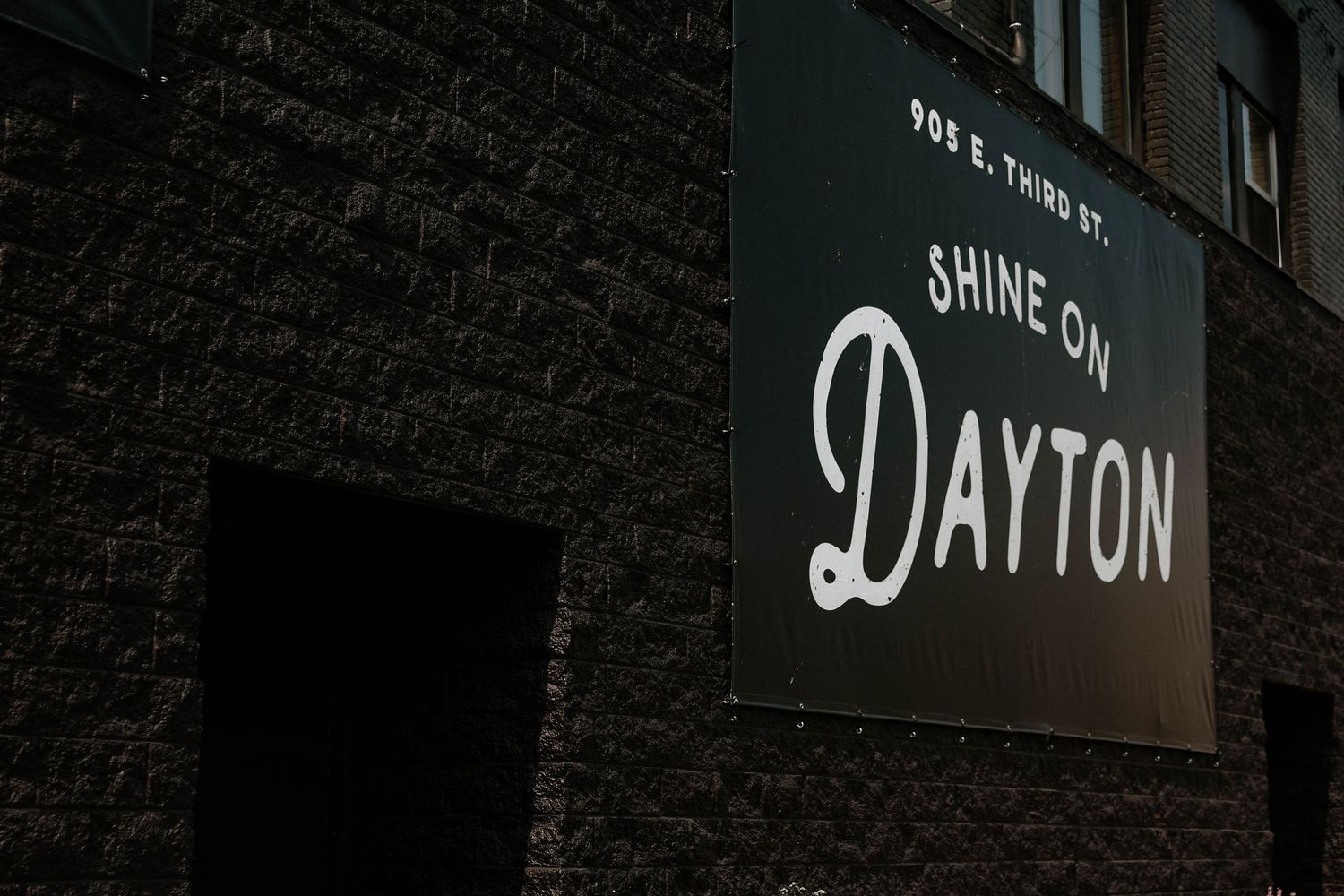 A dark brick building exterior featuring a bold white sign reading Shine On Dayton at 905 E. Third St.