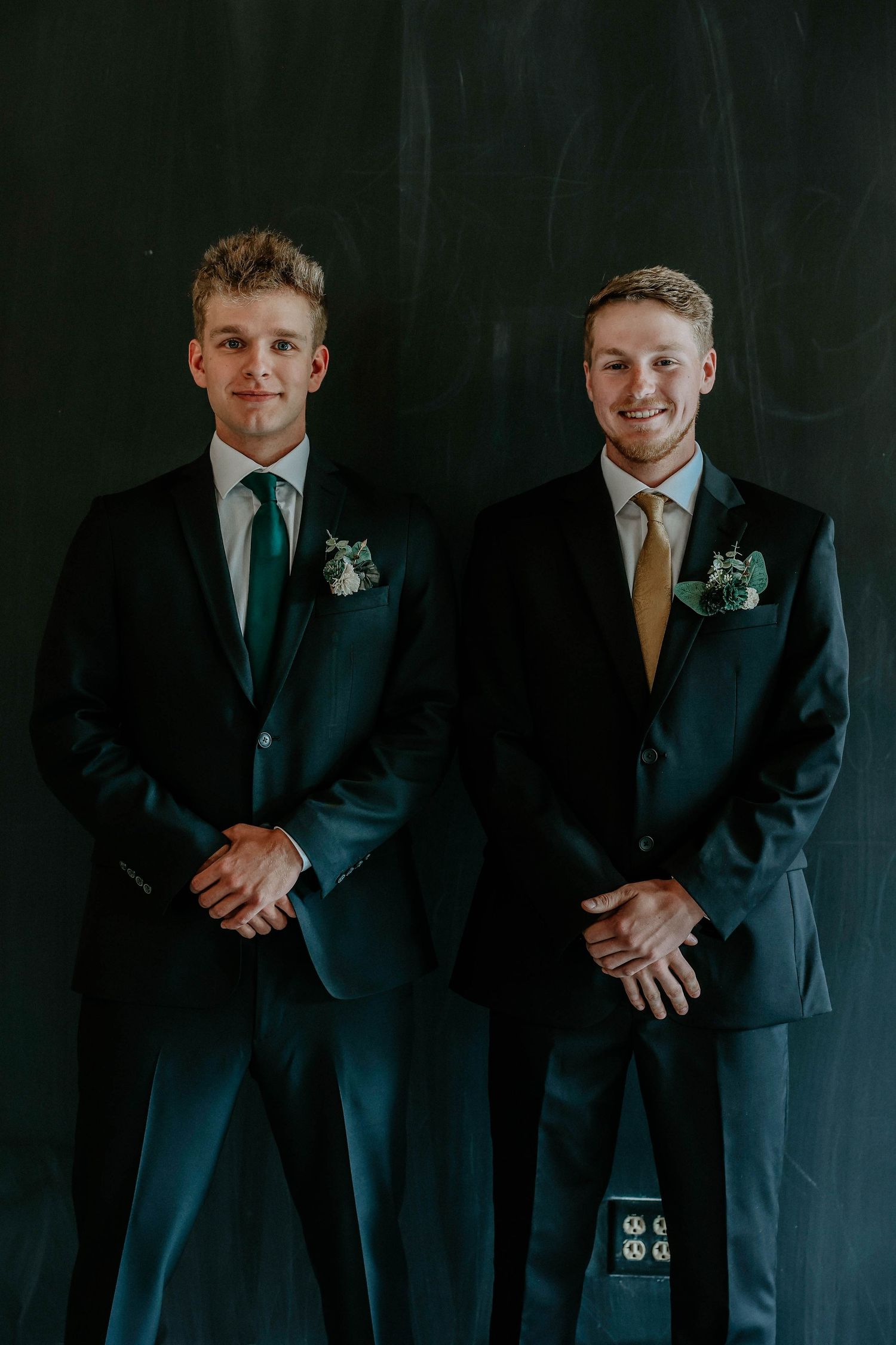 Two young men in dark suits with boutonnieres and ties pose formally against a dark wall.