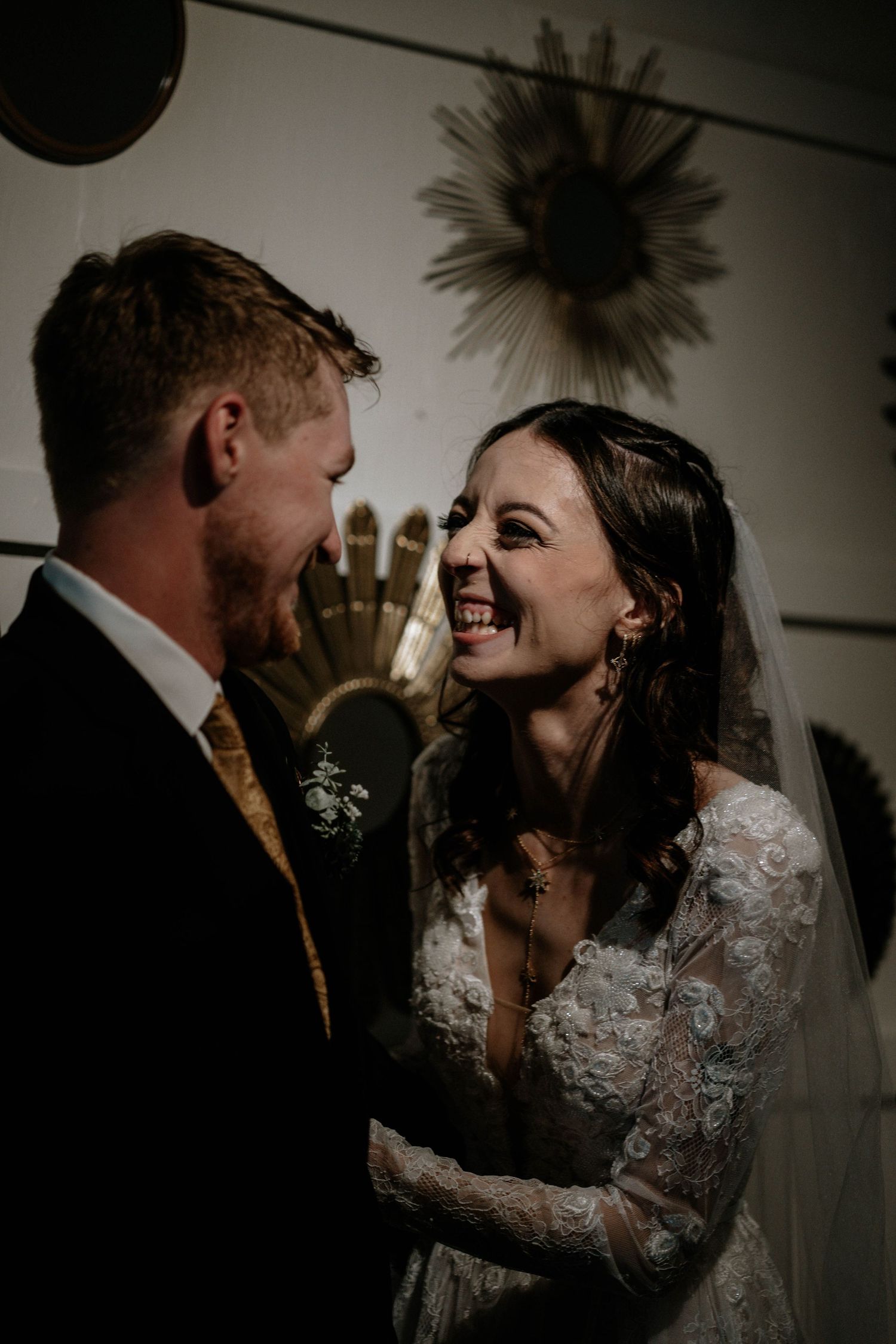 Bride and groom share a joyful moment, smiling at each other in moody wedding portrait with decorative wall art.