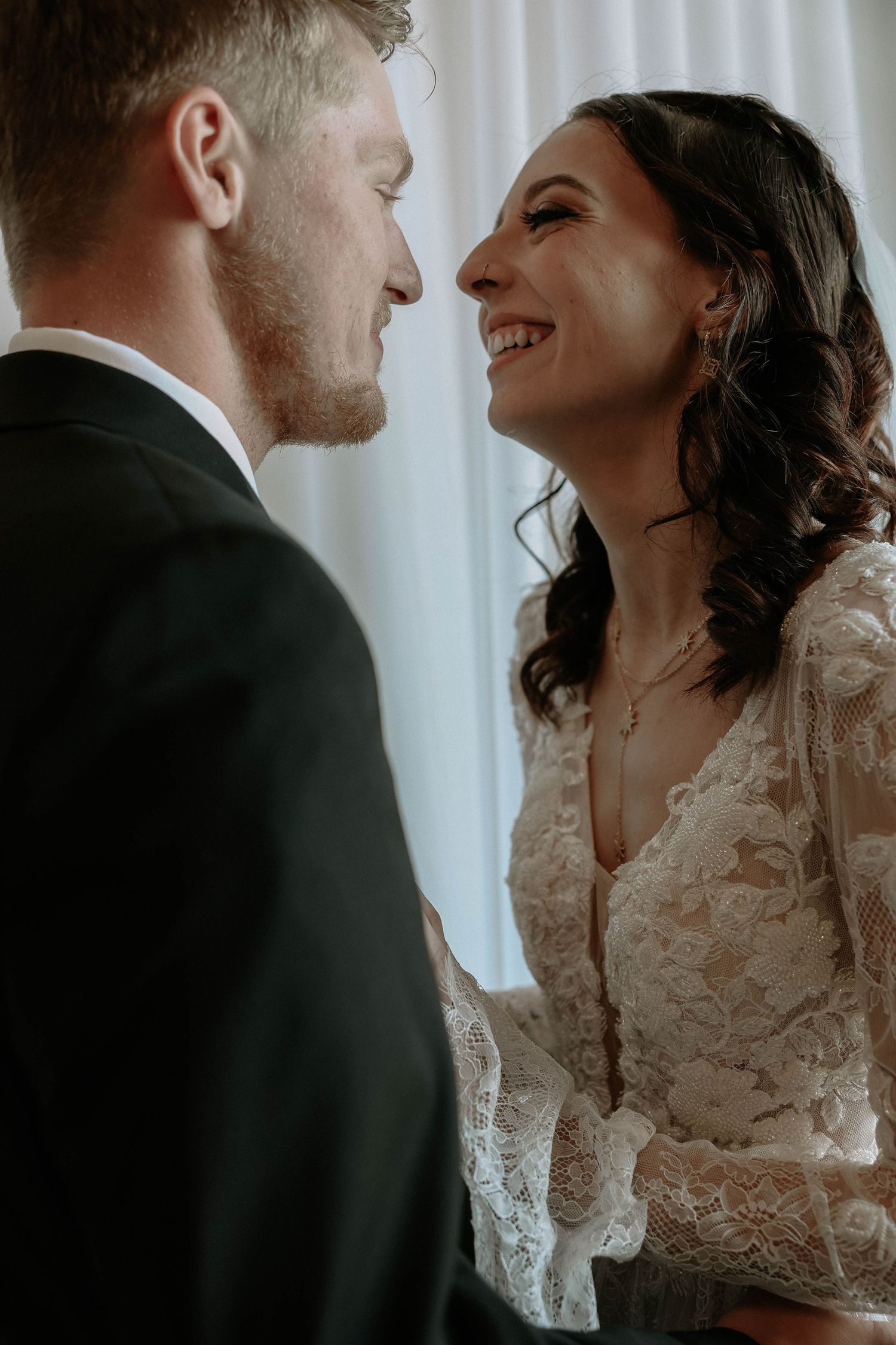 A smiling bride in lace dress shares an intimate moment with her groom in a suit on their wedding day.