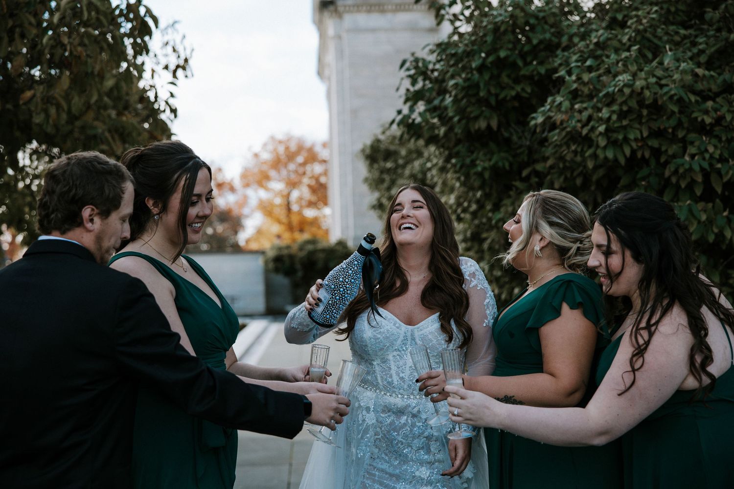 Bride in blue gown pops champagne with bridesmaids in emerald green dresses outdoors at wedding celebration.