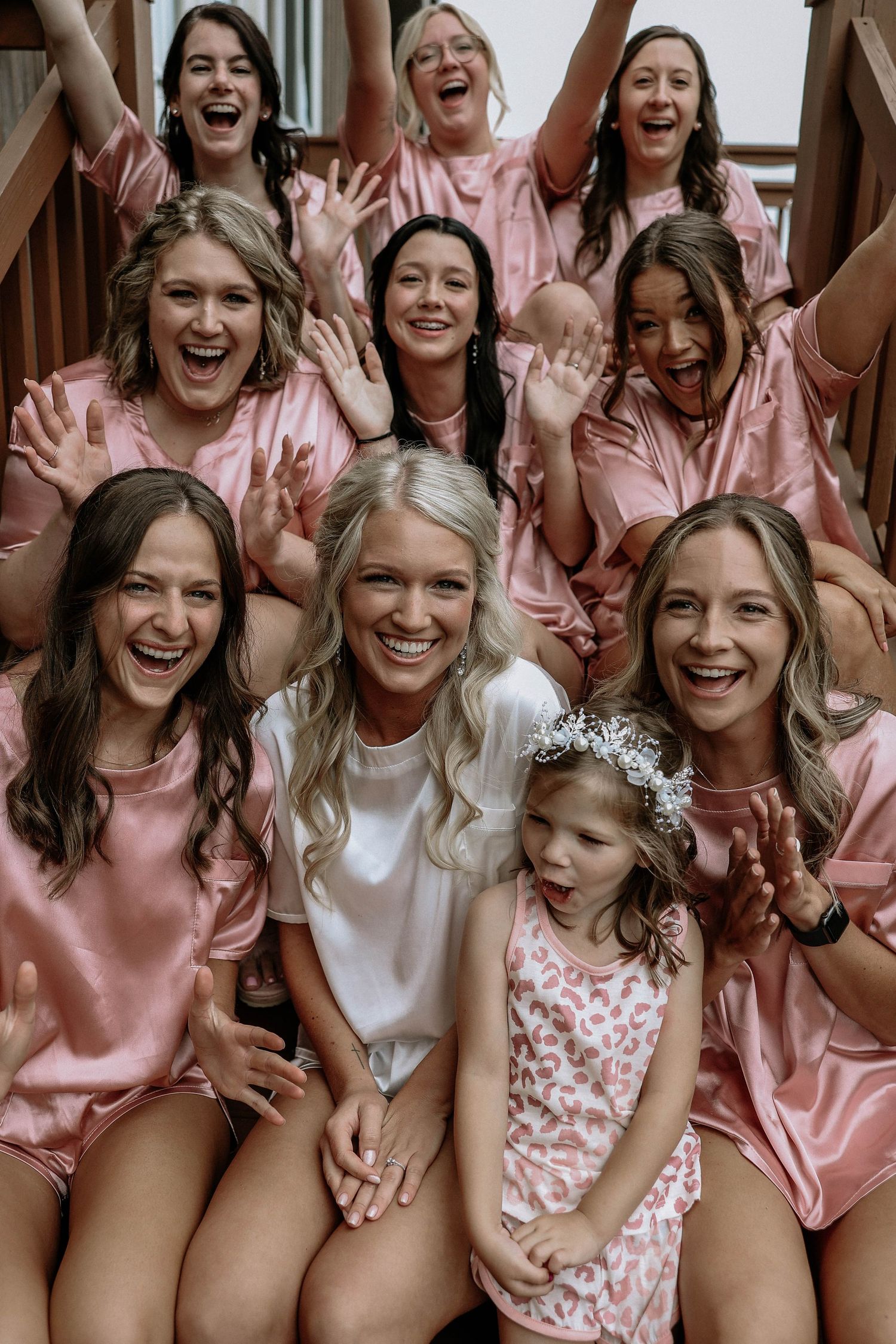 Bride in white with bridesmaids in pink satin robes celebrating together on wooden stairs.