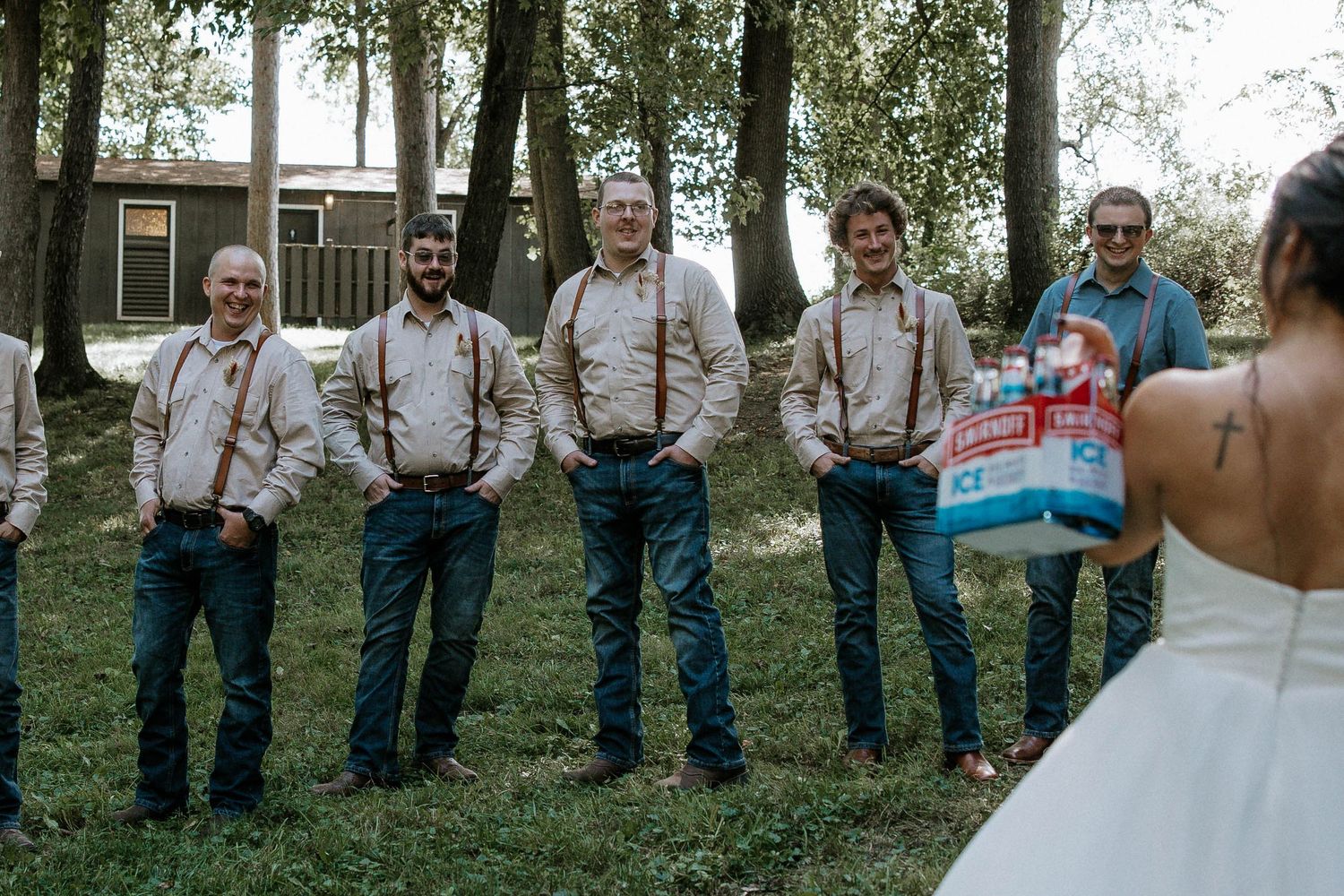 Groomsmen in jeans and suspenders stand outdoors at a rustic wedding, one holding a cooler, facing the bride.