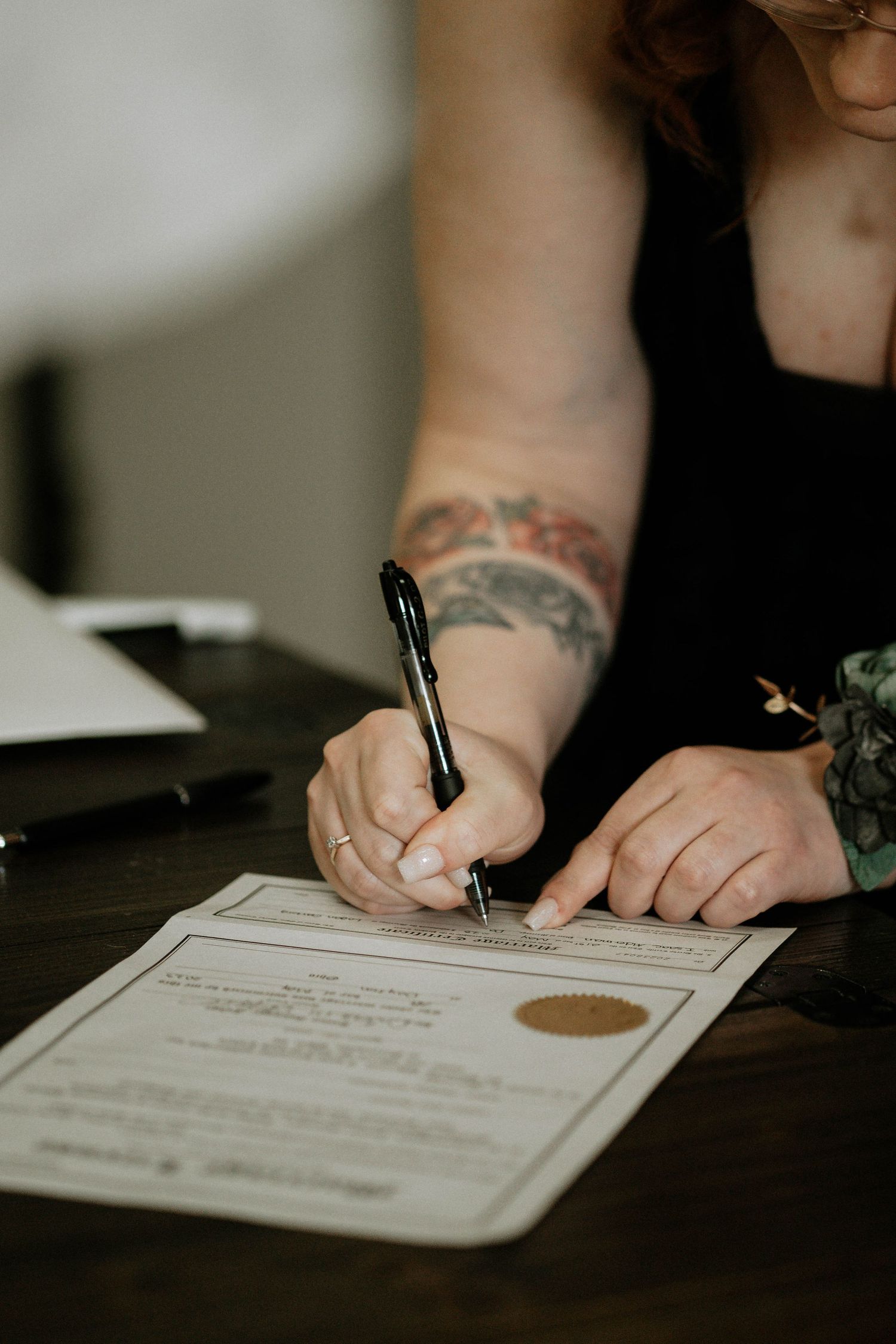 Woman with floral arm tattoo signing an official document with a black pen at a wooden desk.