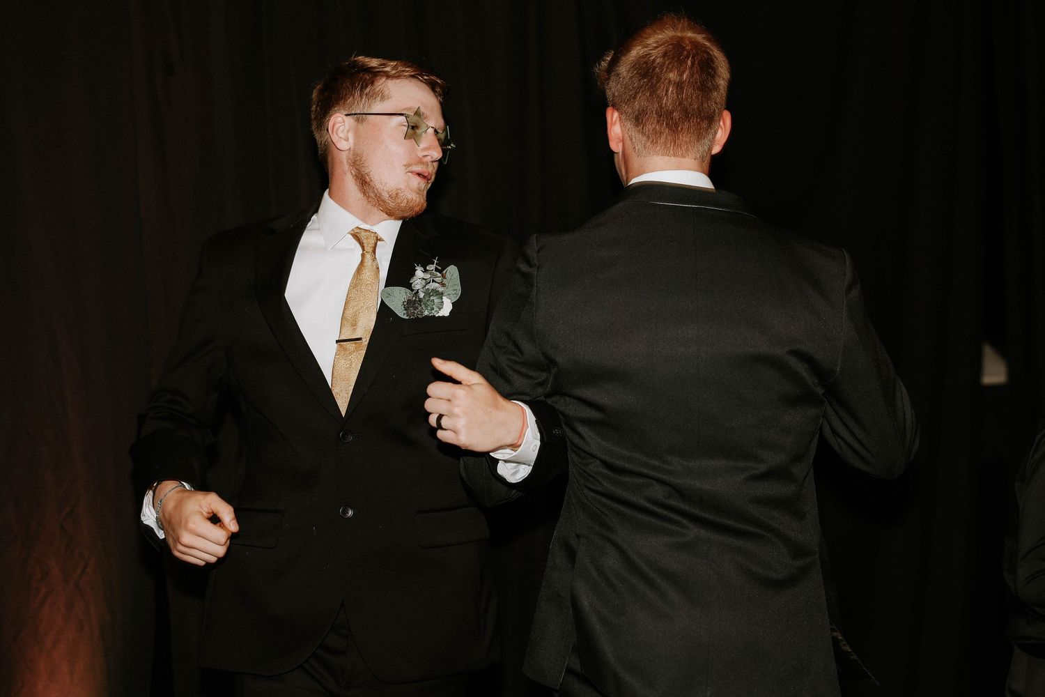 Two men in black suits with boutonnieres share a moment at a formal wedding reception.