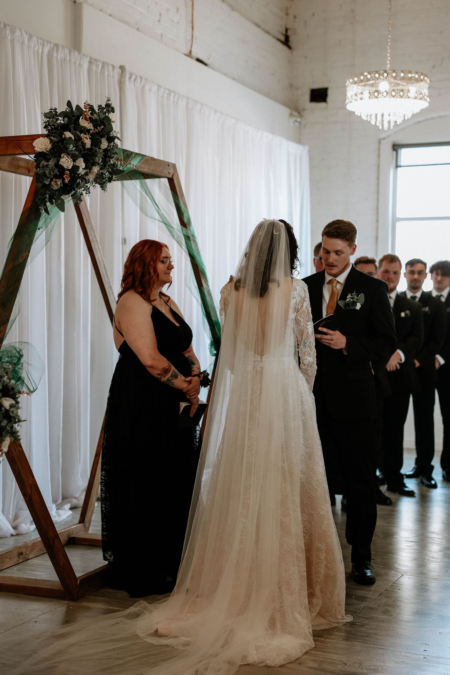 Bride and groom exchange vows at rustic wooden arch altar with officiant in black dress at elegant indoor wedding.