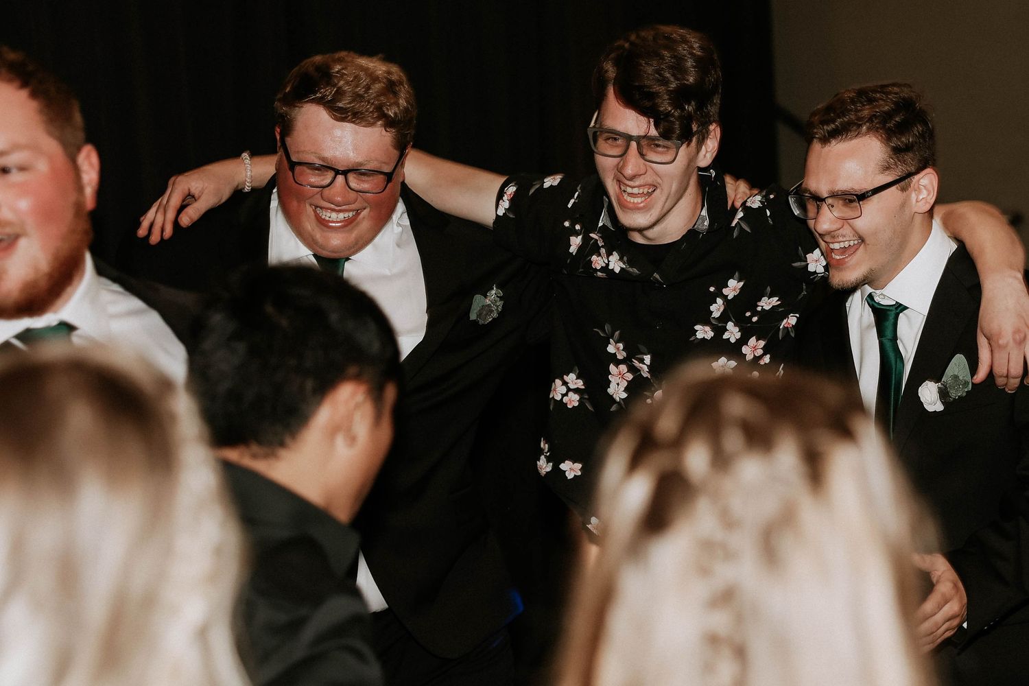 Three young men in formal attire laugh and embrace joyfully at a wedding reception celebration.