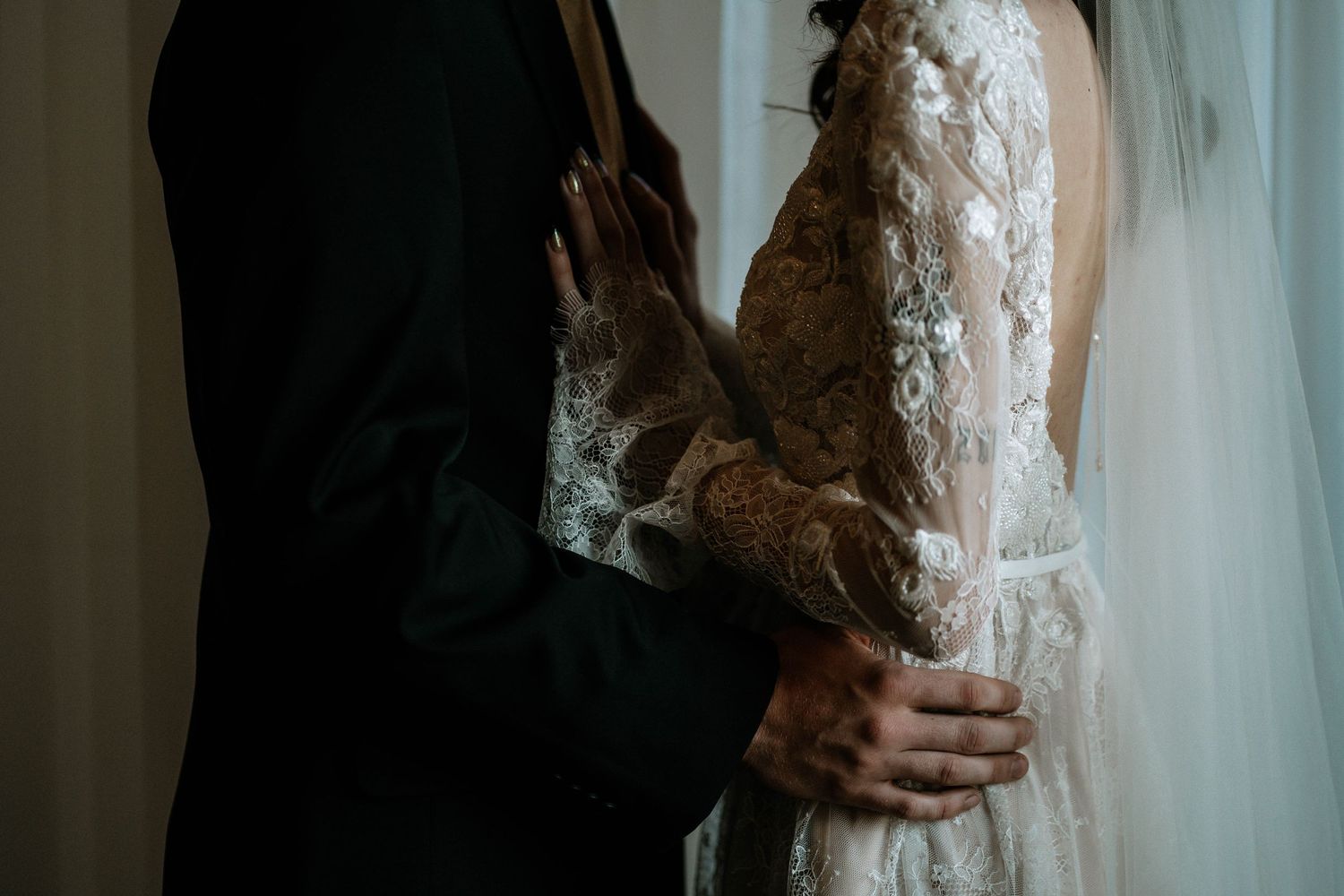 Bride and groom embracing, showcasing intricate lace wedding dress with floral details and elegant veil.