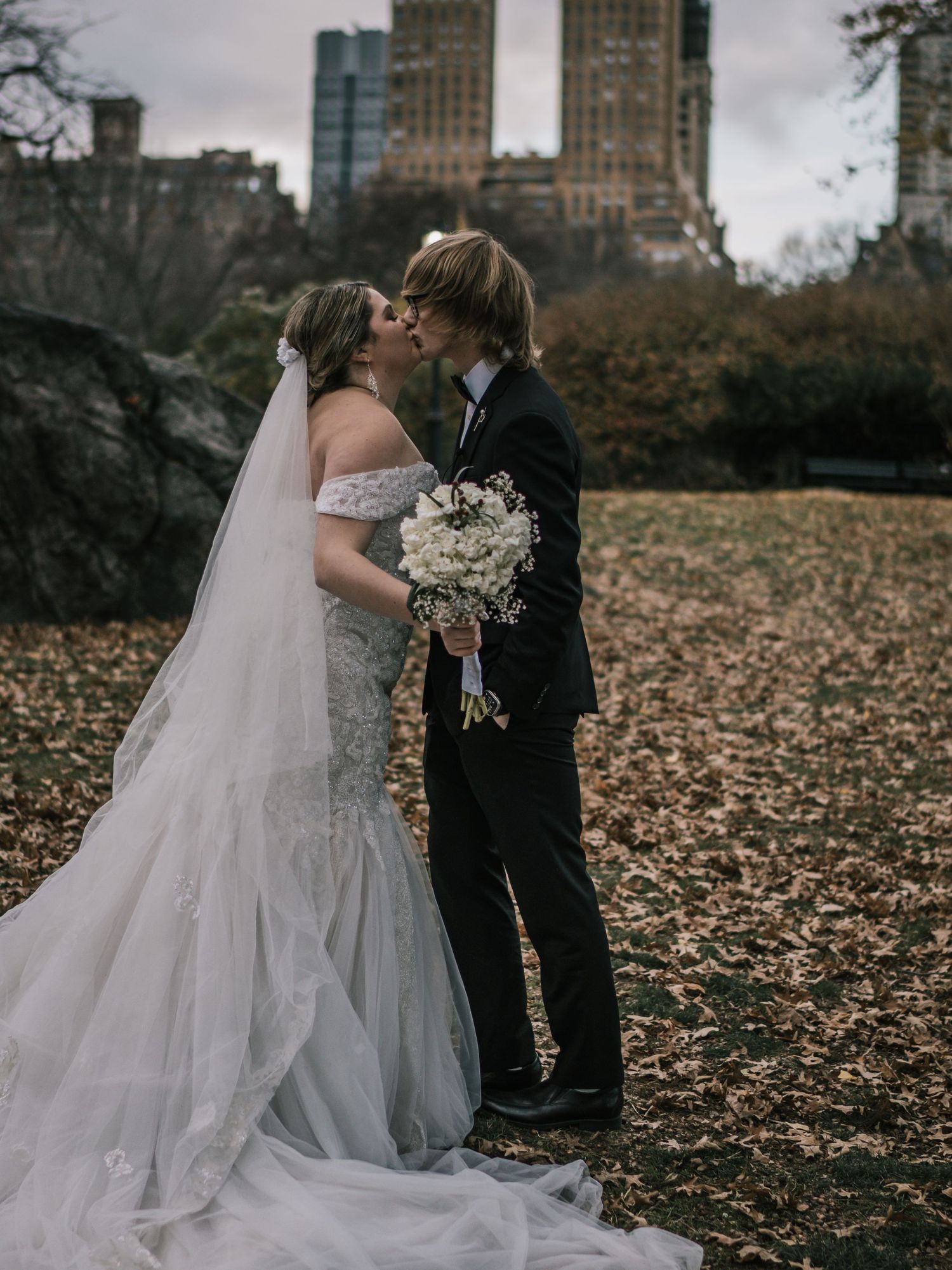 Newlyweds share first kiss surrounded by fallen autumn leaves with New York City skyline in Central Park.