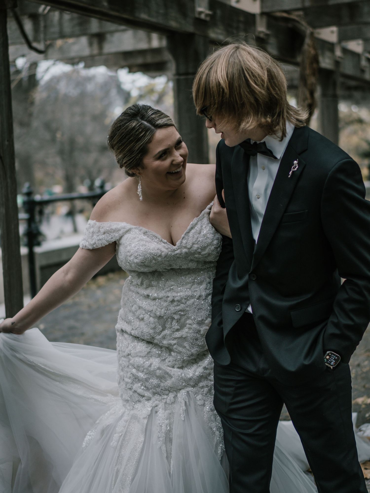 Smiling bride in off-shoulder lace wedding gown twirling skirt while groom in black tuxedo looks on adoringly.