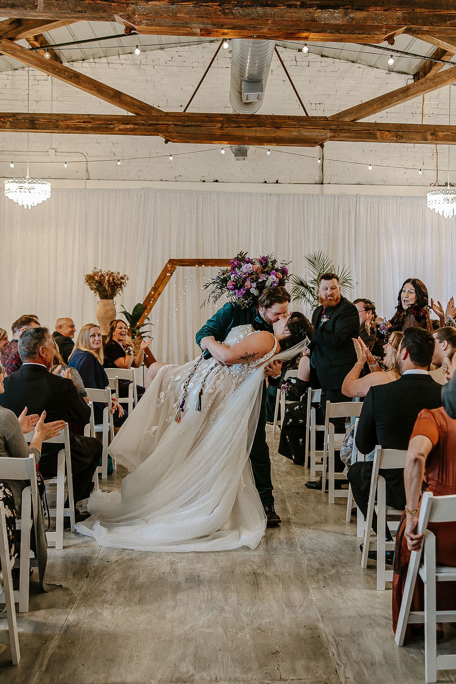 Groom dips bride for romantic kiss in wedding aisle as guests cheer and applaud around them.