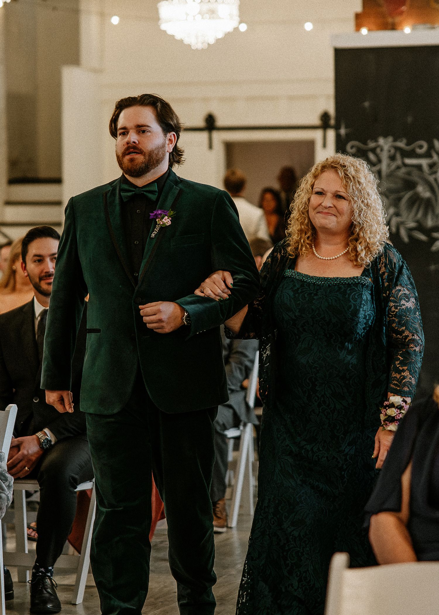 Groom in green velvet suit walks down aisle with his mother in teal lace dress.