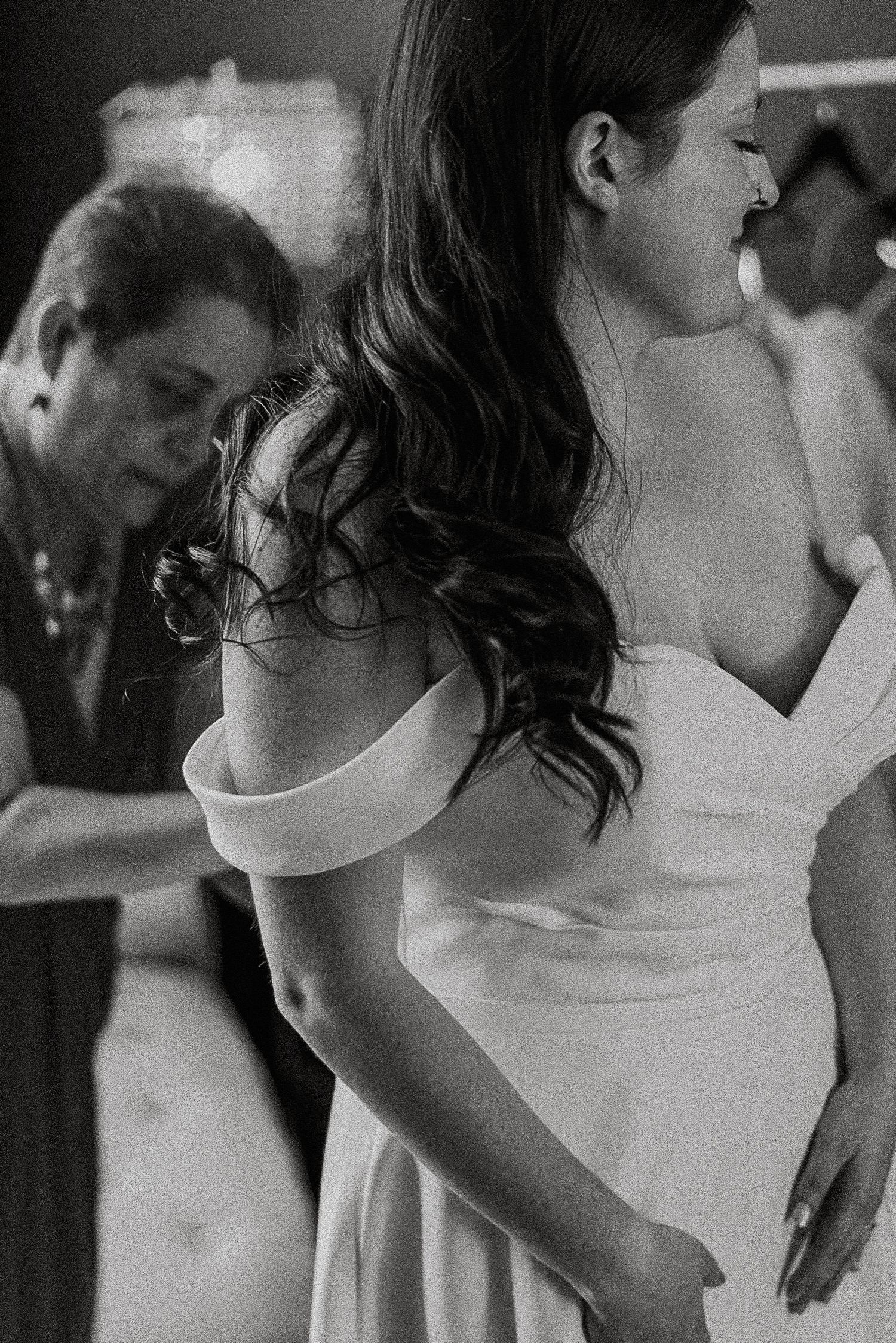 Black and white photo of mother buttoning bride into her elegant off-shoulder wedding dress.