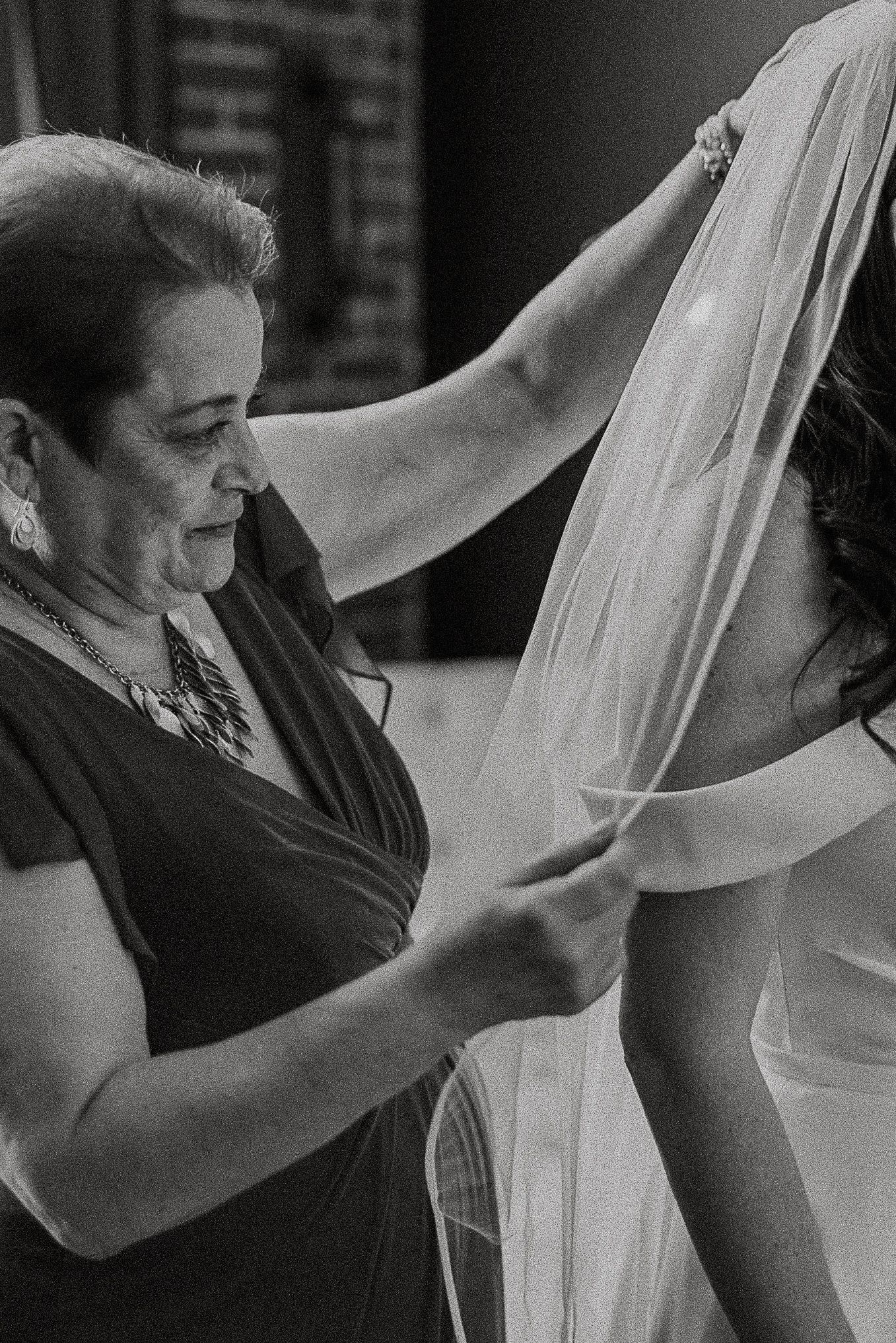 Black and white photo of mother carefully placing veil on bride in wedding gown.