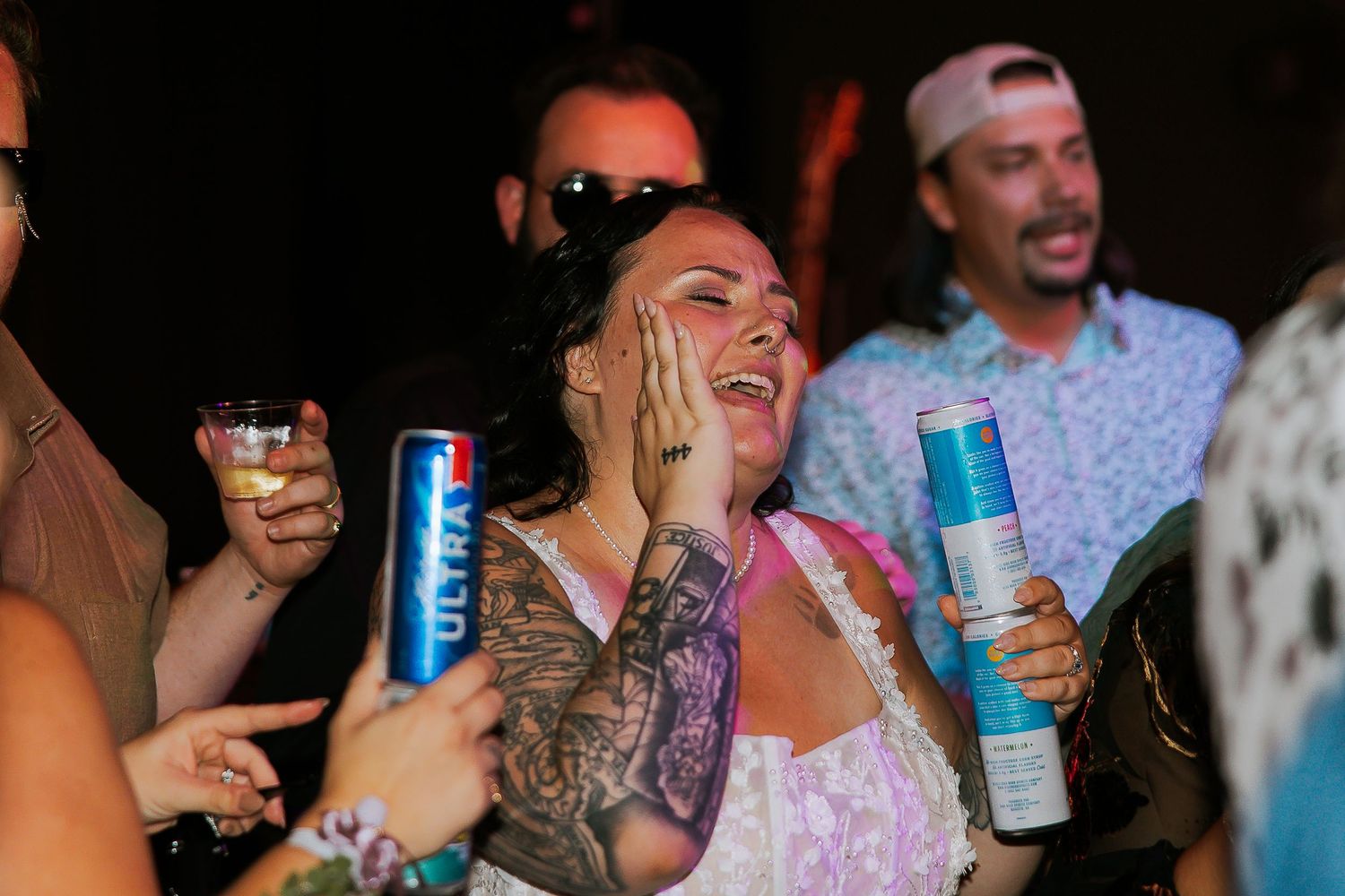 Tattooed woman laughing and holding a Michelob Ultra beer can at a lively outdoor party.