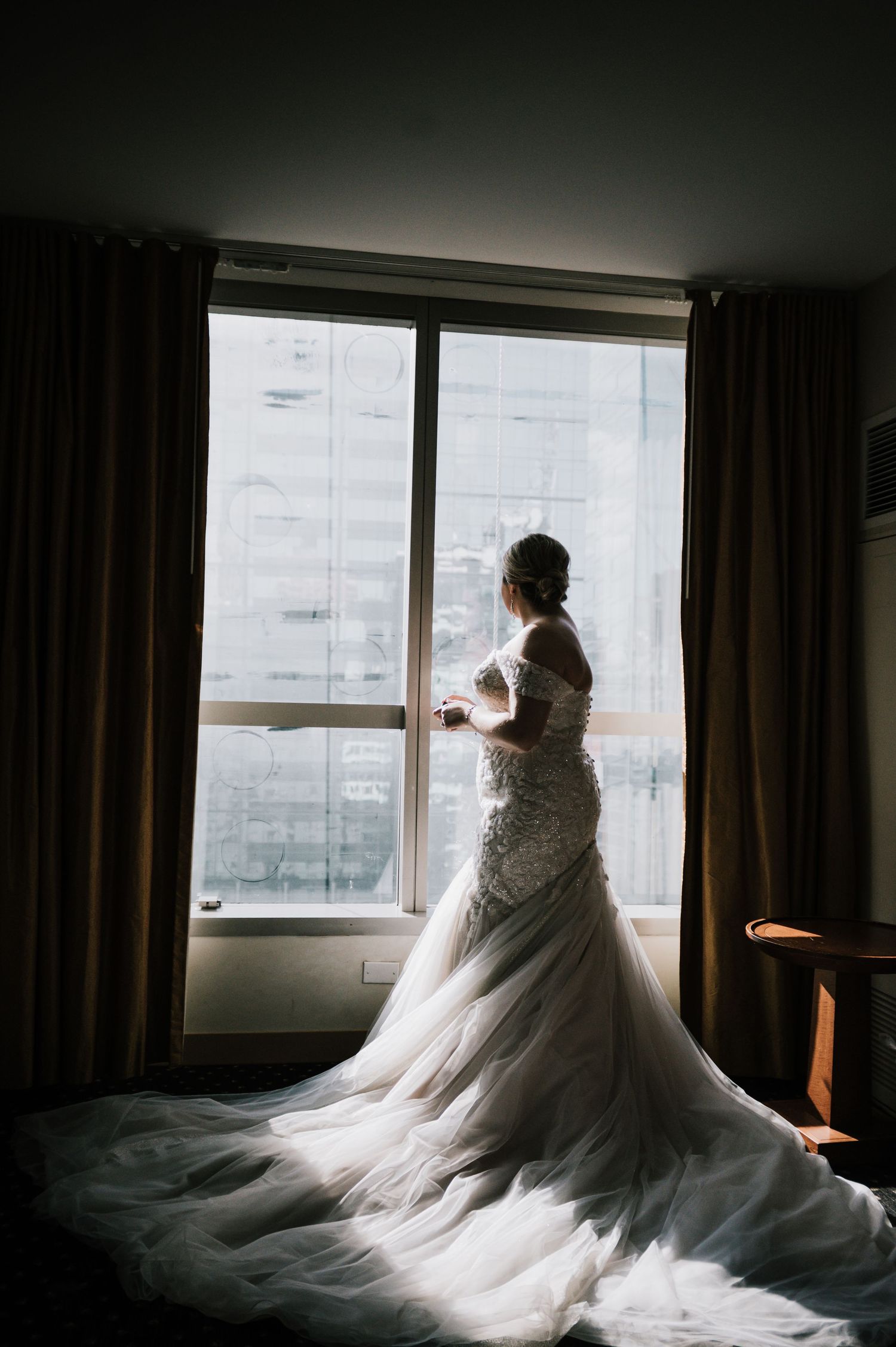 Bride in elegant lace mermaid gown with long train standing by hotel window.