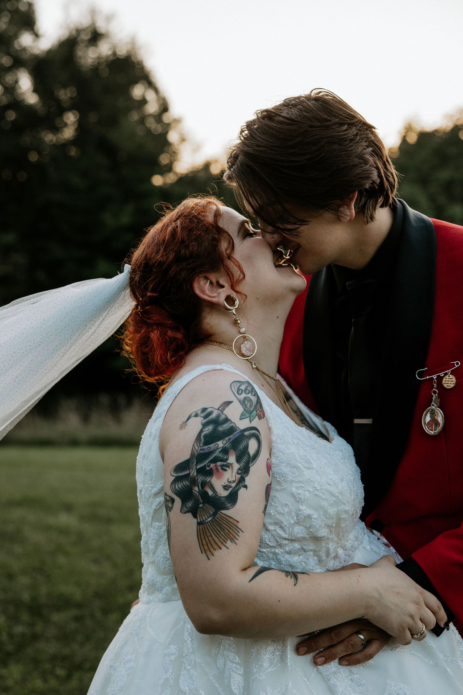 Tattooed bride with red hair shares a kiss with groom in a red jacket at golden hour at the Bella Amore on Enchanted Acres in Dennison, Ohio