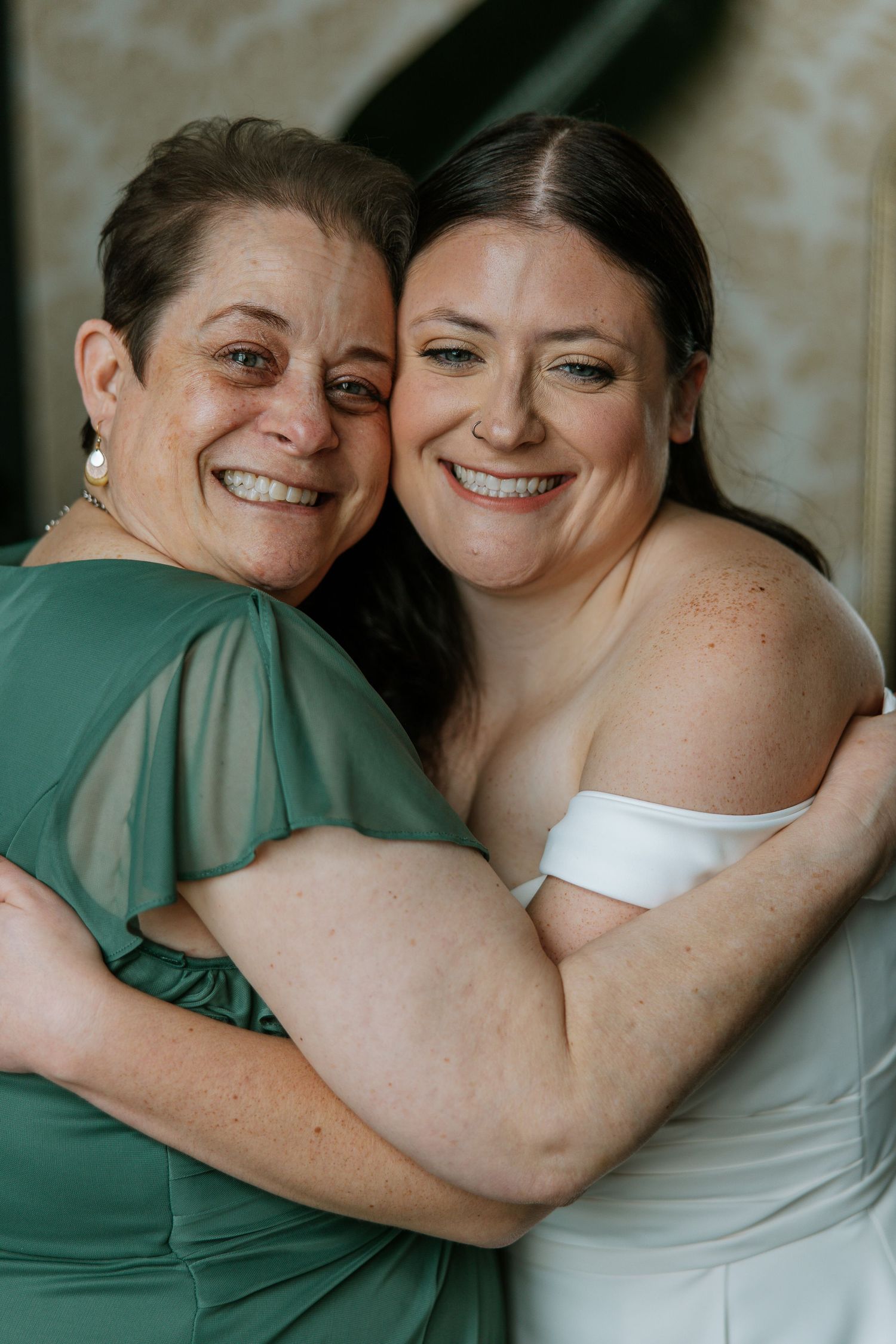 Bride and mother share a joyful embrace, bride in white off-shoulder gown and mother in elegant green dress.
