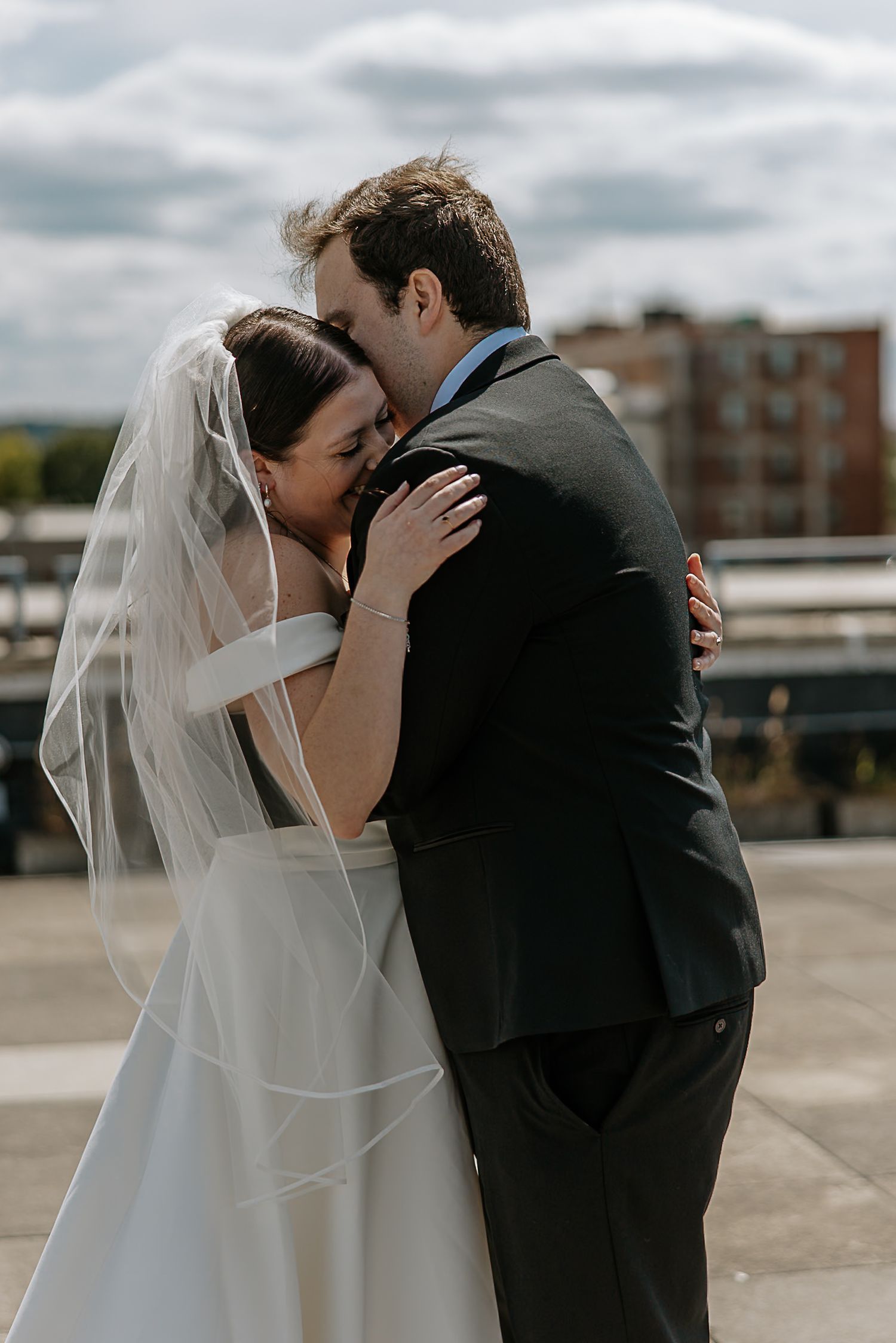 Bride and groom share tender embrace on rooftop, bride in white gown and veil, groom in dark suit.