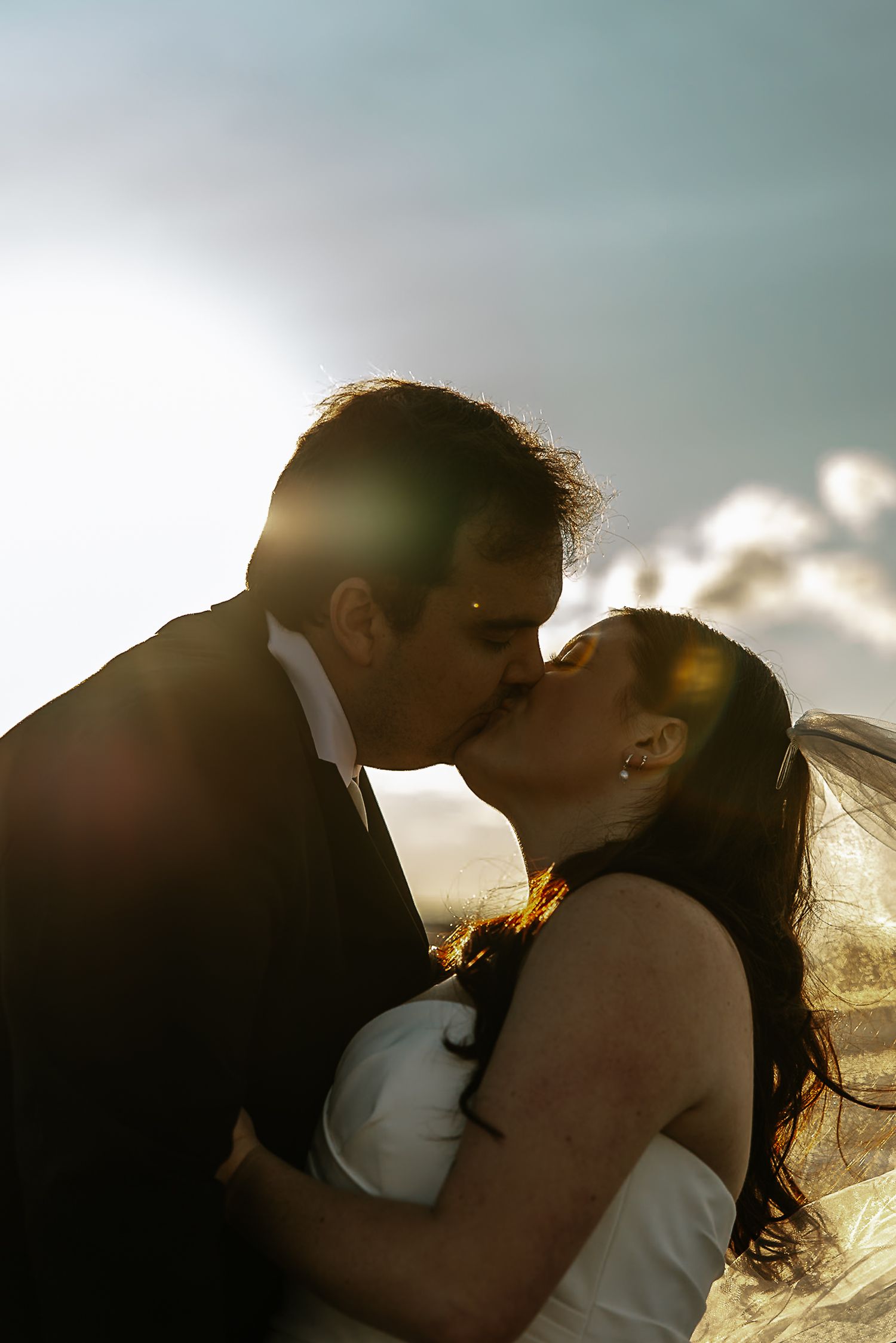 Bride and groom kissing in silhouette against bright blue sky with veil flowing.