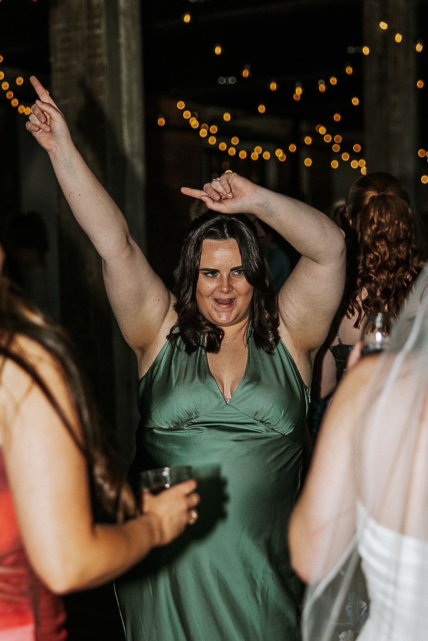 A bridesmaid in a green satin dress dances energetically with arms raised at a wedding reception.