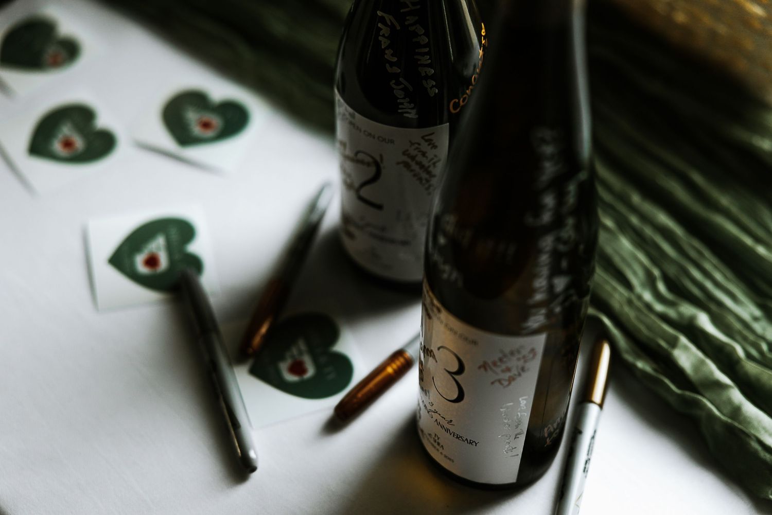 Wine bottles with heart-shaped cards and metallic pens displayed on a white table at a wedding.