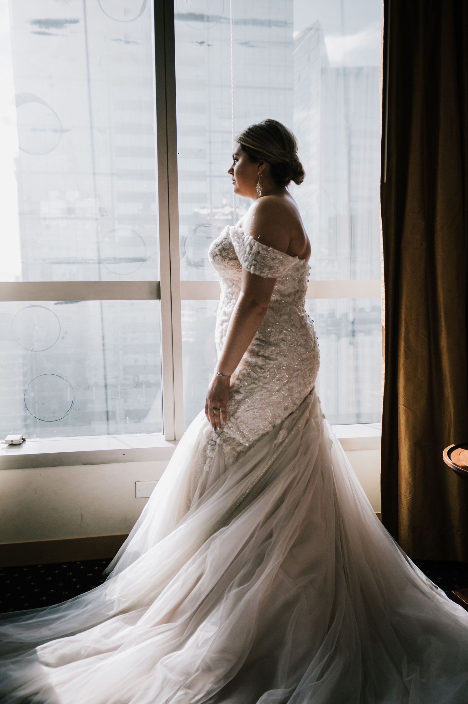 Bride in elegant off-shoulder beaded mermaid gown with long train stands by sunlit city window.