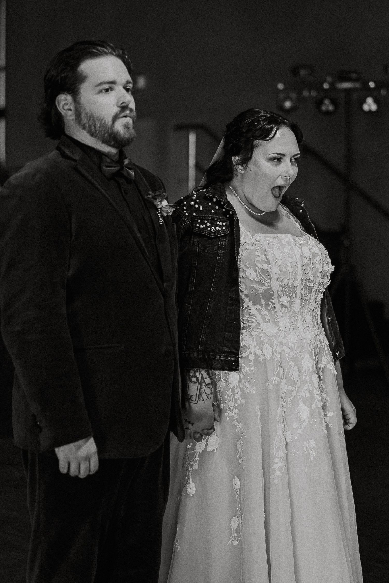 Black and white photo of bride in lace gown and leather jacket with groom looking surprised.