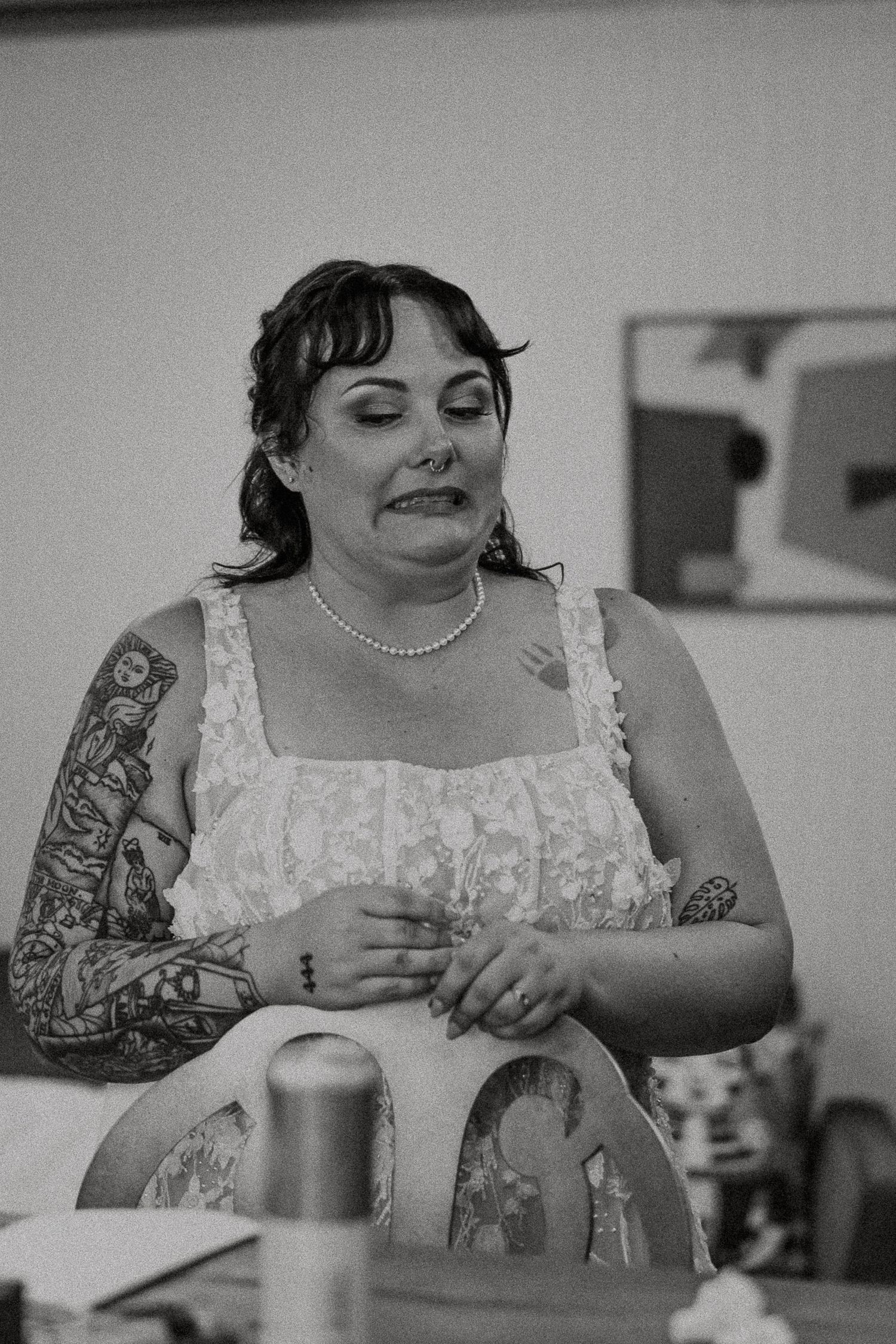 Emotional tattooed bride in white lace dress wearing pearl necklace in black and white photo.