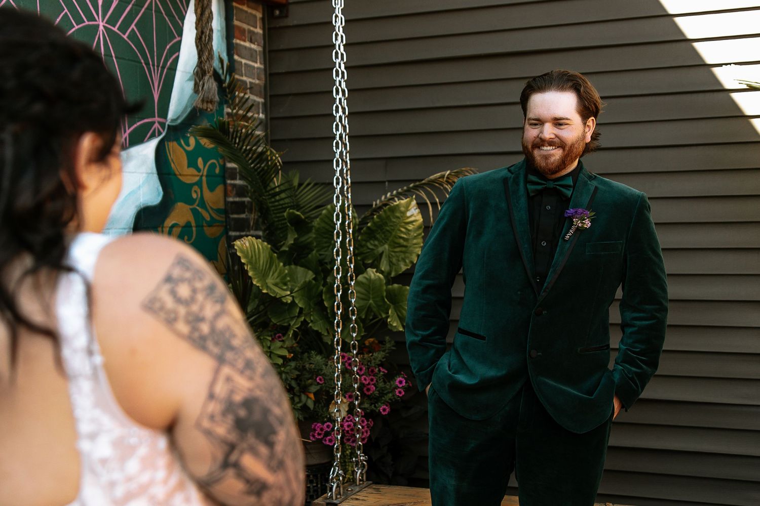 Groom in dark green velvet suit smiling joyfully at tattooed bride during outdoor wedding first look moment.