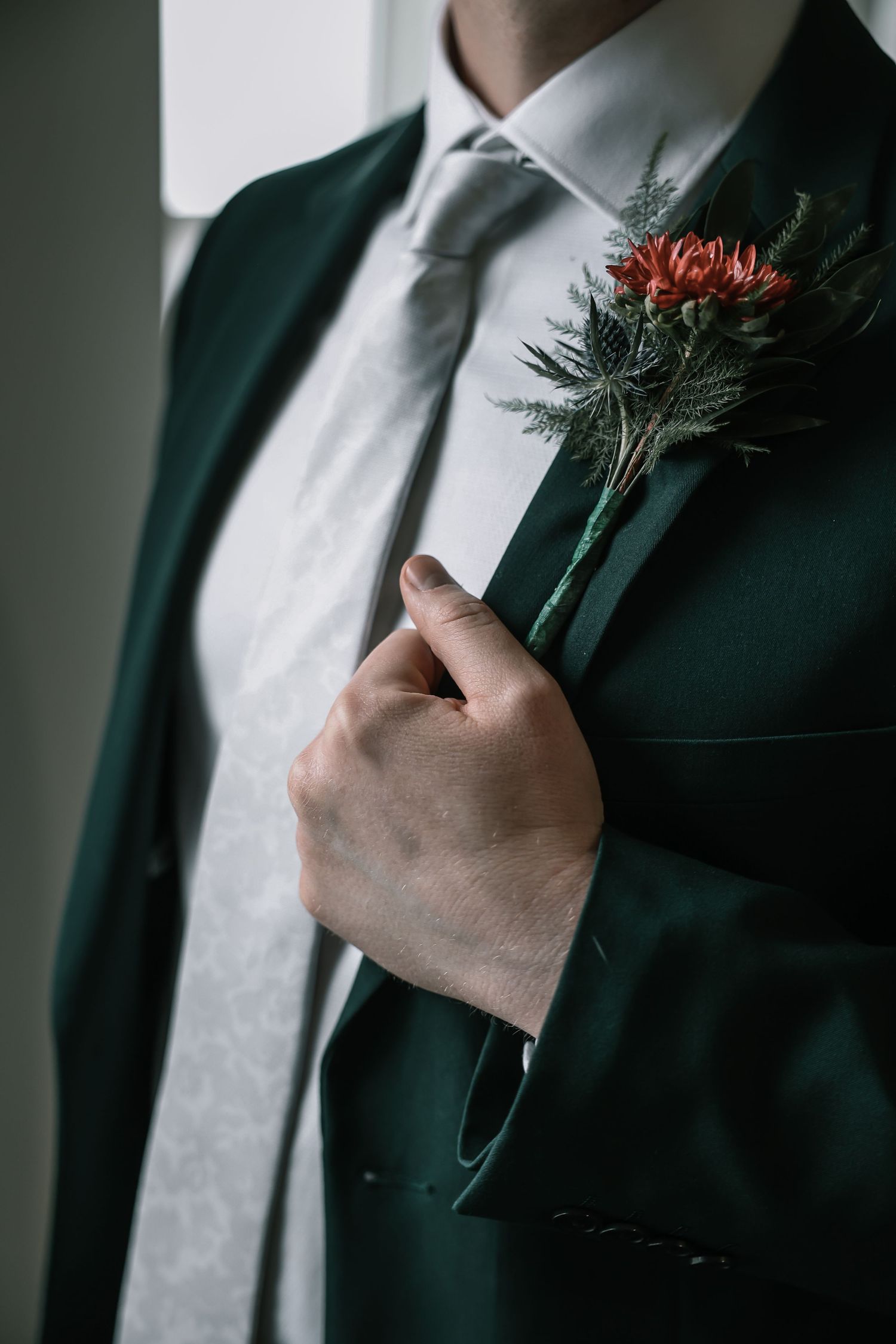 Groom in dark green suit adjusting boutonniere with red flower and greenery accents at The Vue in Columbus, Ohio