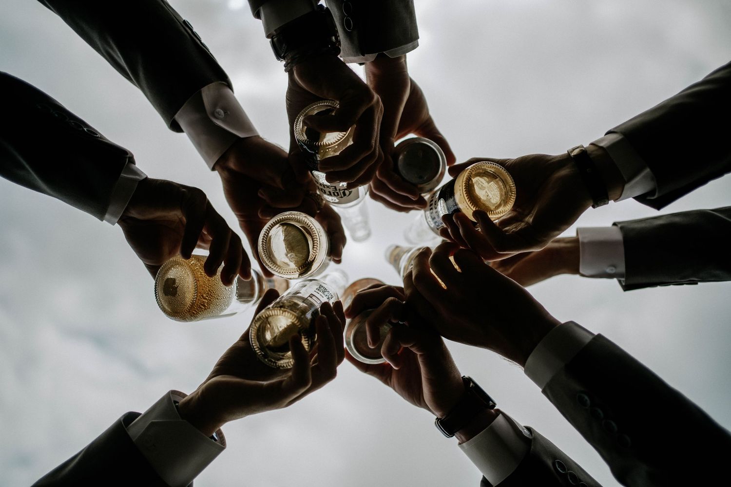Wedding party toasts champagne glasses in a circle huddle photographed from below against cloudy sky.