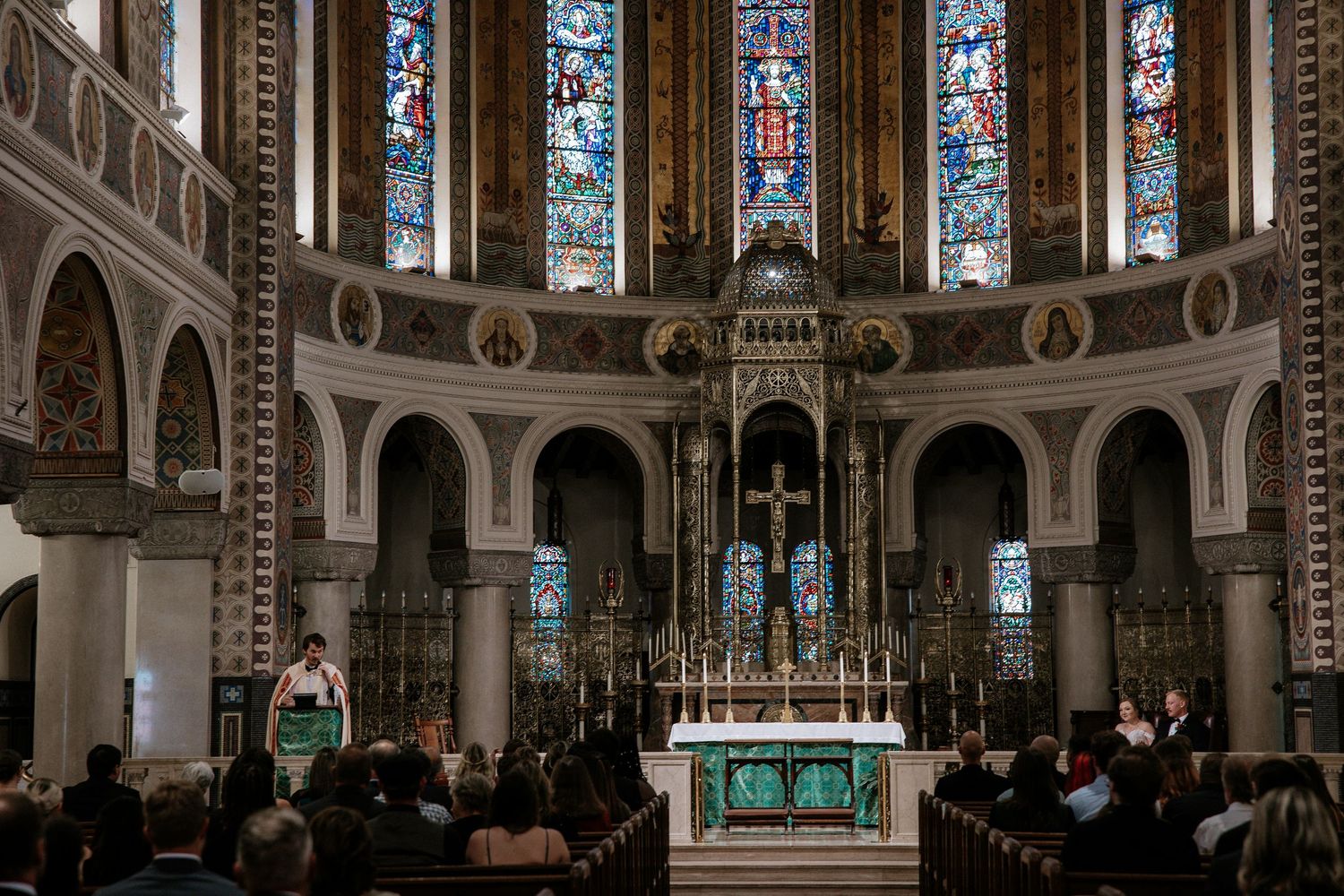 Wedding guests seated in church pews as officiant speaks at elaborately decorated altar with stained glass in Cincinnati, Ohio