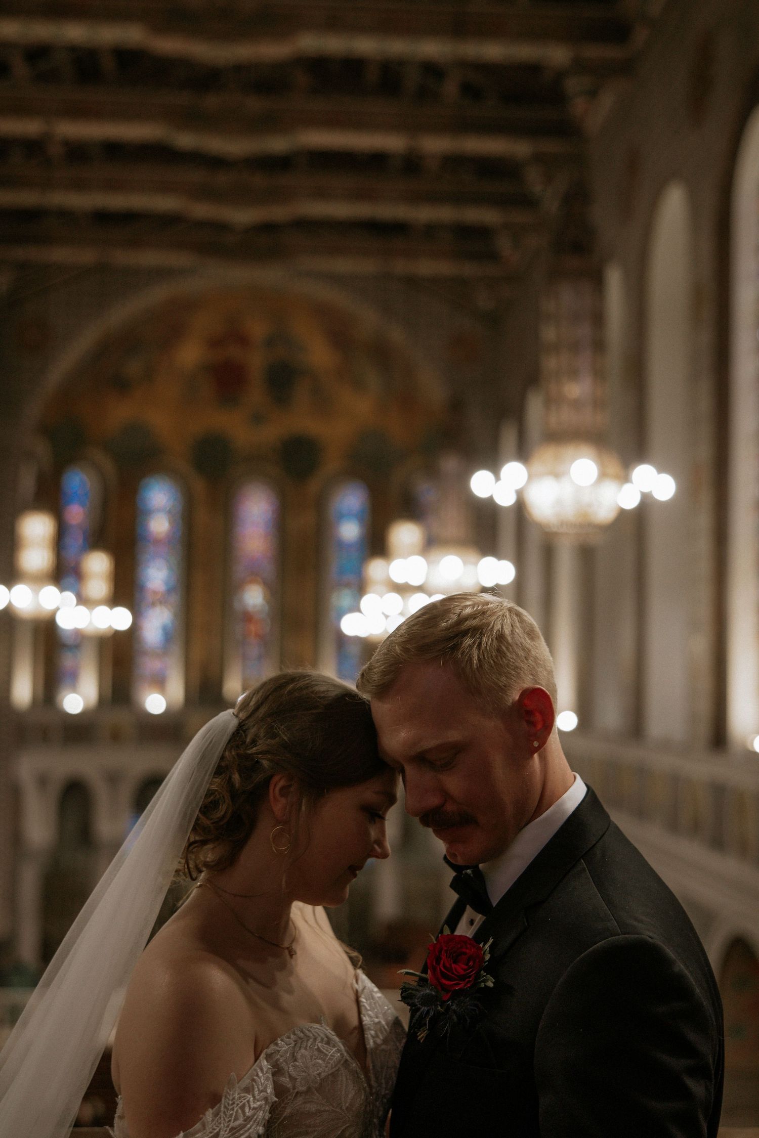 Intimate portrait of newlyweds with foreheads touching beneath ornate cathedral ceiling and chandelier lighting in Cincinnati, Ohio