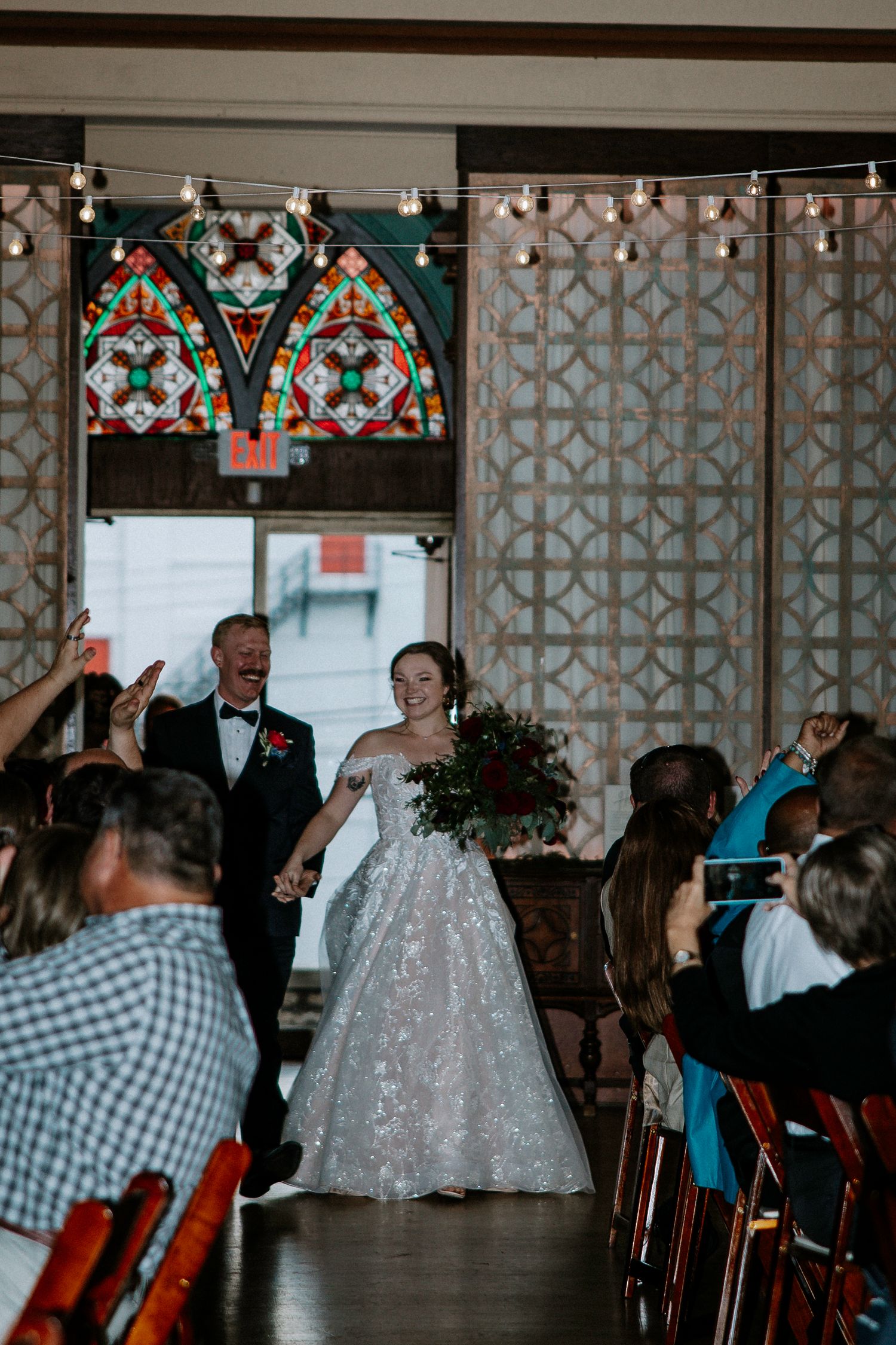 Joyful bride and groom walk down the aisle in a venue with stained glass windows at Urban Artifact in Cincinnati, Ohio